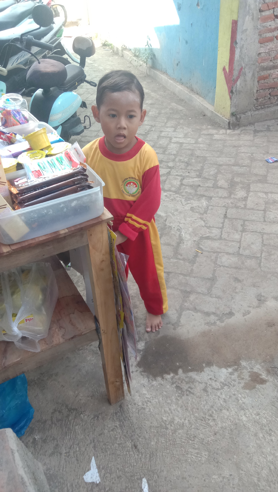 A young boy with short, dark hair and an open mouth stands next to a wooden stall. He is wearing a red and yellow long-sleeved shirt with a circular emblem on the chest and matching red pants with a yellow stripe down the side. The stall has a top shelf filled with various packaged snacks, including what appear to be chocolate bars and small yellow containers. A clear plastic bin holds some of these items. Below the top shelf, there is another shelf with plastic bags, one of which contains a white substance and has the word "FU" visible. A blue plastic bag is on the ground near the stall. The ground is paved with concrete that appears wet in some areas. In the background, a motorcycle is partially visible, and a brick wall with blue and yellow painted sections runs along the side. The scene is likely an outdoor market or roadside stall in Karang Anyar, Indonesia.