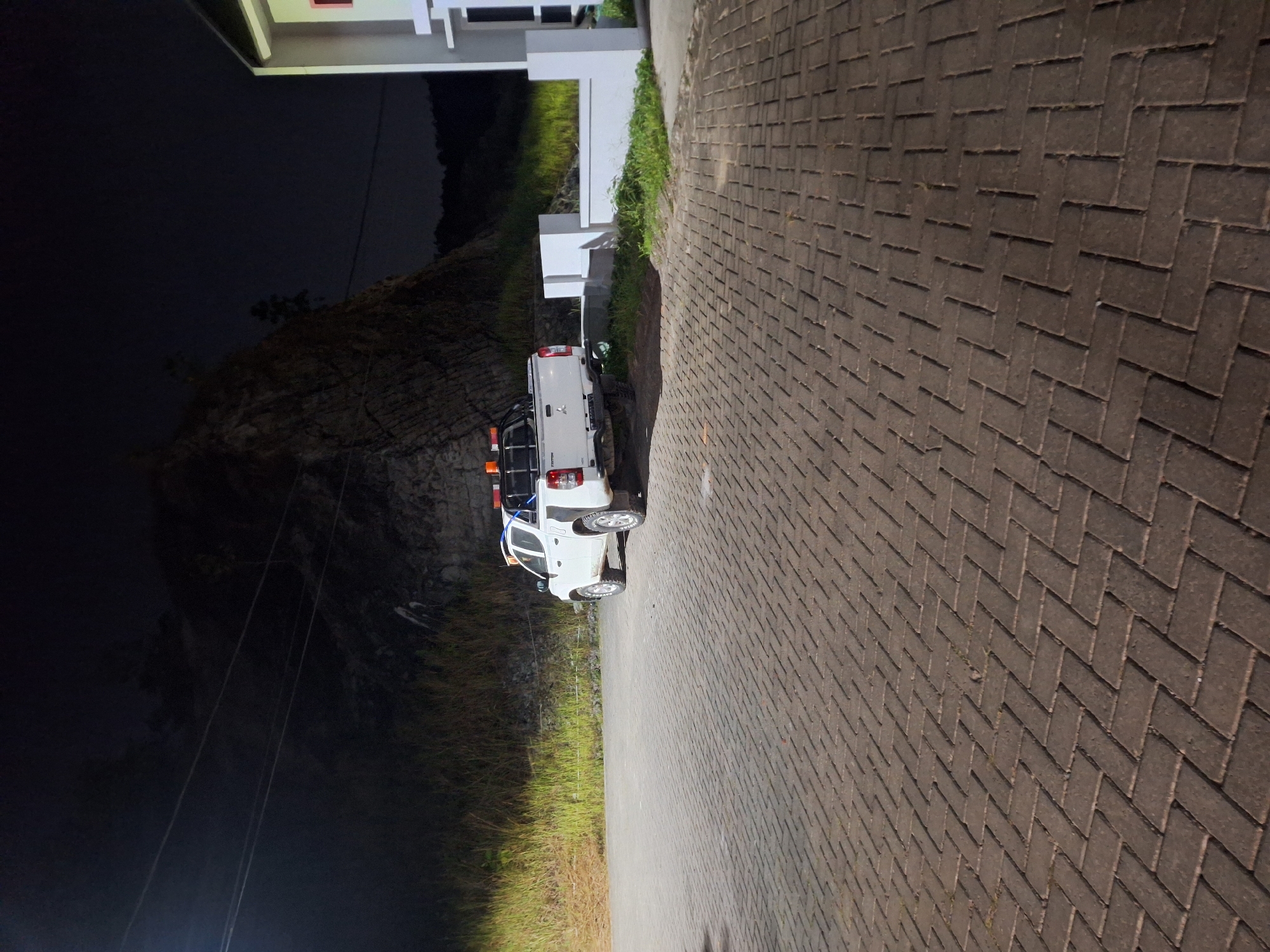 The image depicts a nighttime scene in a paved outdoor area. A white pickup truck, possibly a Mitsubishi Triton, is parked in the center of the frame. The truck has emergency lights mounted on its roof and bed, suggesting it may be a utility or service vehicle. Behind the truck, a steep, rocky embankment rises into darkness. To the right of the truck, a modern building with white architectural features is partially visible. The foreground is dominated by a textured paving of rectangular bricks laid in a herringbone pattern. The lighting is artificial, casting shadows and illuminating the immediate area around the truck and building, while the background fades into darkness. The scene appears to be a parking lot or an access road adjacent to a building and a natural landscape feature. The provided information indicates this scene is located in Samarinda, Indonesia.
