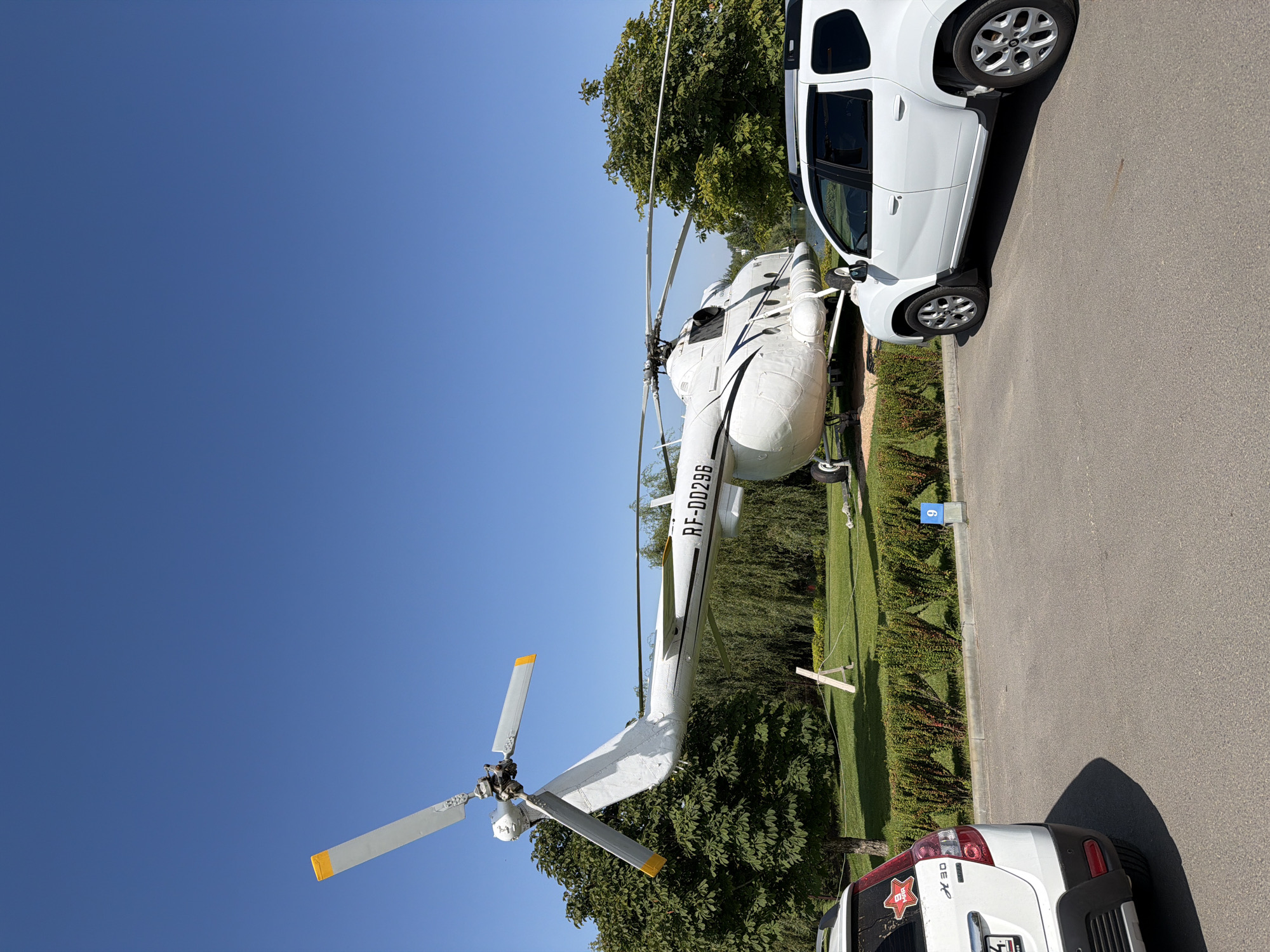 A white helicopter, identified by the registration RF-DD296, is displayed on a grassy area under a clear blue sky. The helicopter has two large main rotor blades, each tipped with orange, and a smaller tail rotor. It is situated in a park-like setting with mature trees and manicured bushes. In the foreground, two white vehicles are parked on an asphalt surface. The vehicle on the left is a white hatchback with a red star decal on the rear windshield. The vehicle on the right is a white SUV. A sign with the number "9" is visible in the background, near the bushes. The scene is likely in Kamyshin, Russia, suggesting this is an outdoor display or museum.