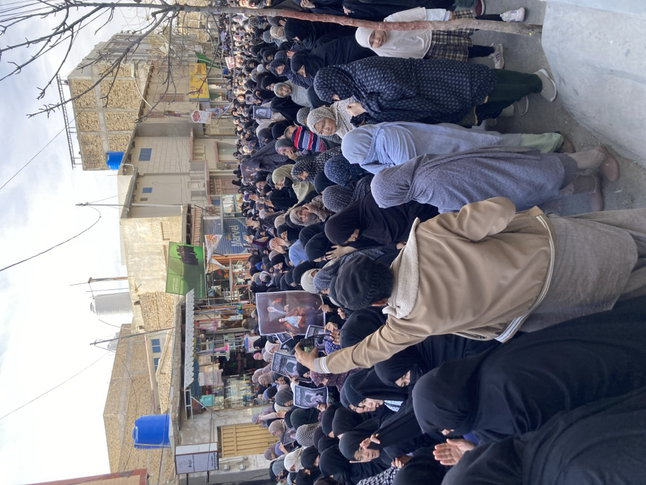 The image captures a large crowd of people gathered on an urban street in Quetta City Tehsil, Pakistan, during daylight. The majority of attendees are women, many dressed in black abayas or hijabs, while others wear varying colors of traditional head coverings and attire. Men and children are also present within the dense gathering.
In the foreground, a man, viewed from the rear, wearing a beige jacket over a shalwar kameez and a dark cap, extends his right arm upwards, holding a rectangular poster. This poster features a colored portrait of two individuals. Numerous other individuals throughout the crowd are also holding up similar posters, many depicting single portraits. Several people are visible with their hands raised in the air.
The background consists of multi-story buildings with flat roofs, characteristic of South Asian urban architecture. Blue and white cylindrical water tanks are visible on several rooftops. Ground-level storefronts with awnings are present, and overhead electrical wiring crisscrosses the sky. A bare-branched tree is partially visible on the right side of the frame. The overall impression is a public demonstration or gathering.