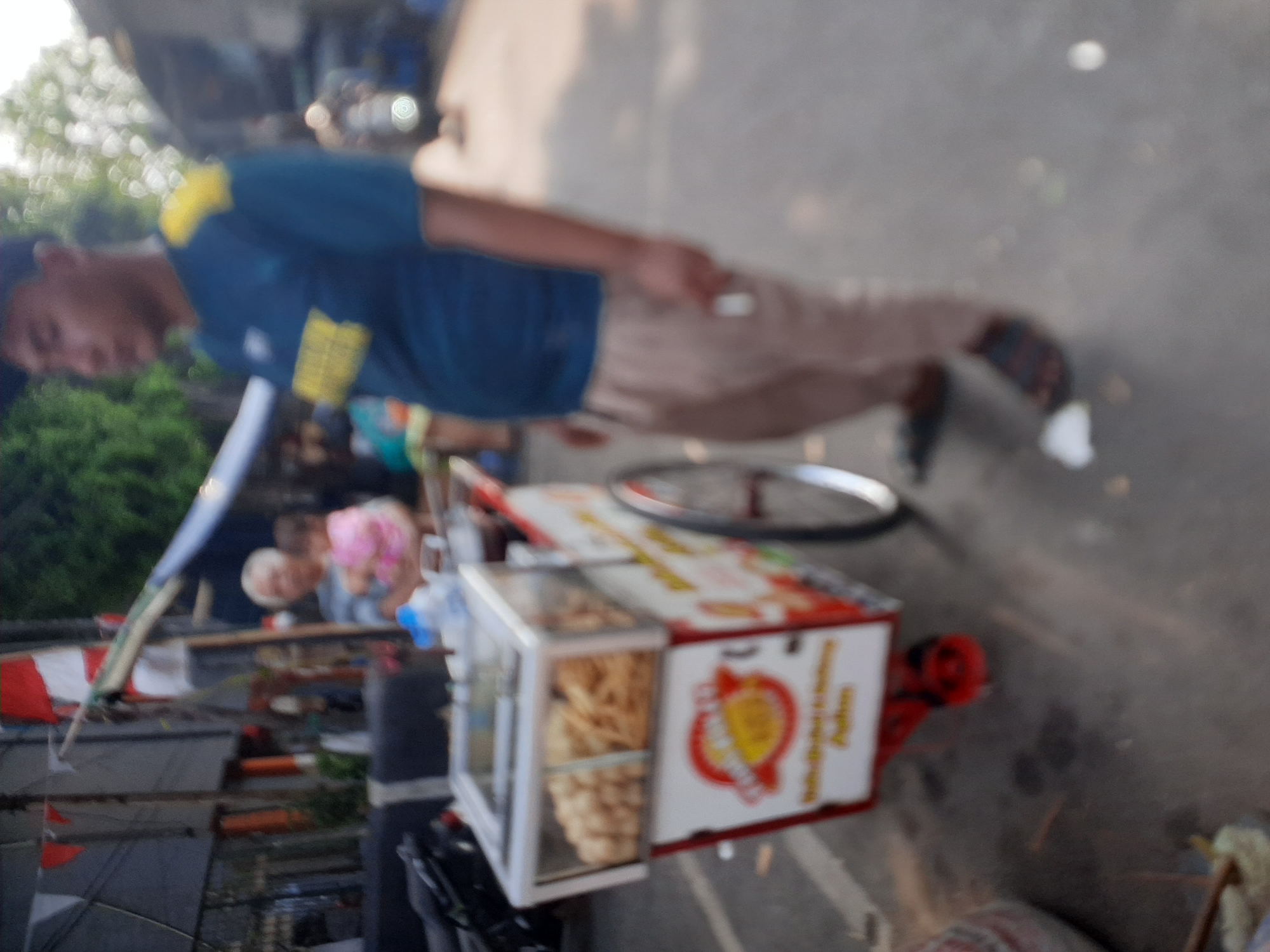 In Central Jakarta, Indonesia, an outdoor scene unfolds featuring a street vendor with a cart. The vendor, a man wearing a blue t-shirt with yellow accents and light-colored pants, stands near his cart. The cart, primarily white and red, displays various food items in a glass-fronted compartment. The cart also features branding with a circular logo and text. In the background, an elderly person holds a baby, and flags are visible. The environment appears to be a street, with trees and buildings in the distance, bathed in natural light.