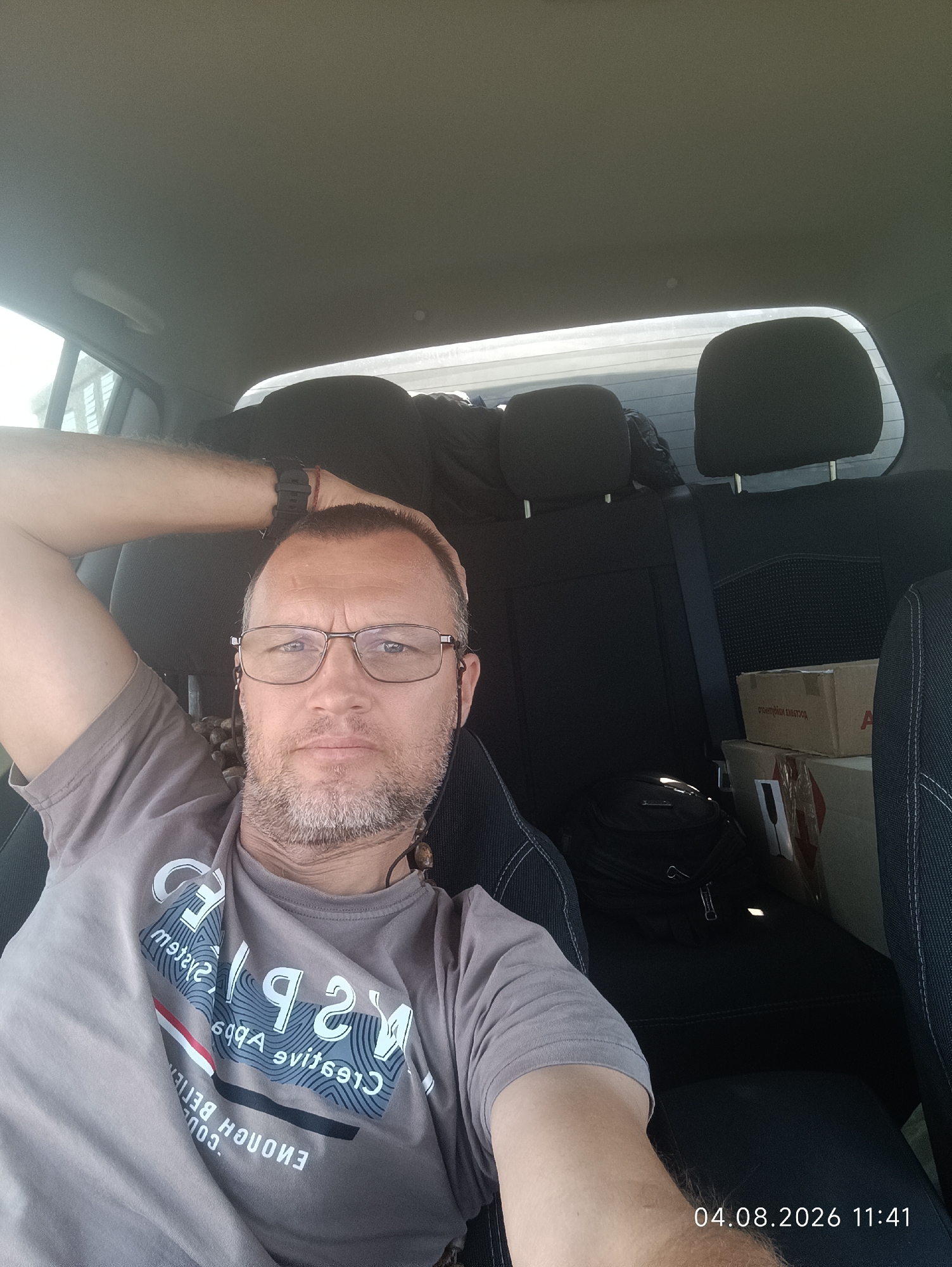 A man in his 40s with short brown hair and a beard, wearing glasses, a grey t-shirt, and a watch, is seated in the back seat of a car. He is looking at the camera with his left arm behind his head. The interior of the car, including the seats and headrests, is visible. There are also boxes and bags in the back seat. The timestamp "04.08.2026 11:41" is visible in the bottom right corner of the image. The location of Starokozache, Ukraine, is inferred from the metadata.