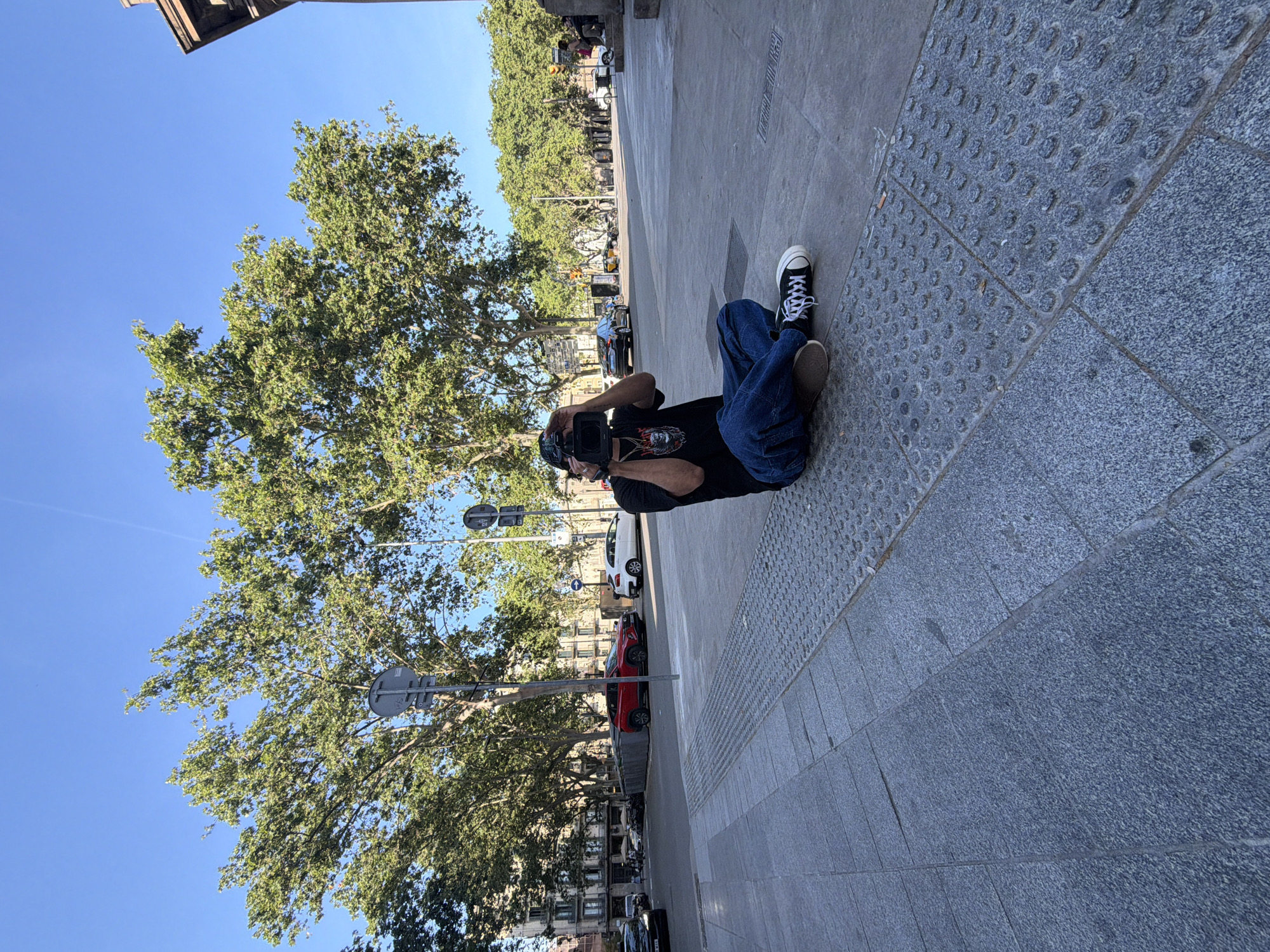 A man lies on his side on a gray stone sidewalk, holding a video camera to his face. He wears blue jeans, a black t-shirt with a graphic design, and black-and-white sneakers. The man appears to be filming or taking a picture. The background includes trees with green foliage, parked cars, street signs, buildings, and a blue sky. The ground near the man's feet features a tactile paving surface with raised dots, indicating a pedestrian crossing for the visually impaired. The location is Barcelona, Spain.