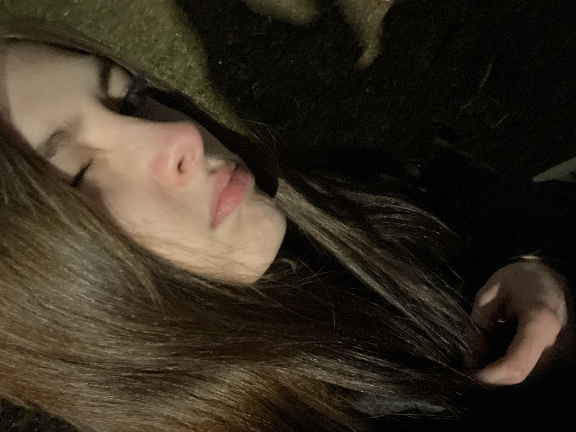A close-up, high-angle shot features a woman with long, brown hair lying down on what appears to be grass. Her eyes are closed, and her facial expression is neutral. Her right hand is visible near her chest. The background is dark, with some vague shapes that suggest foliage or other vegetation. The location cannot be determined based on this image alone.