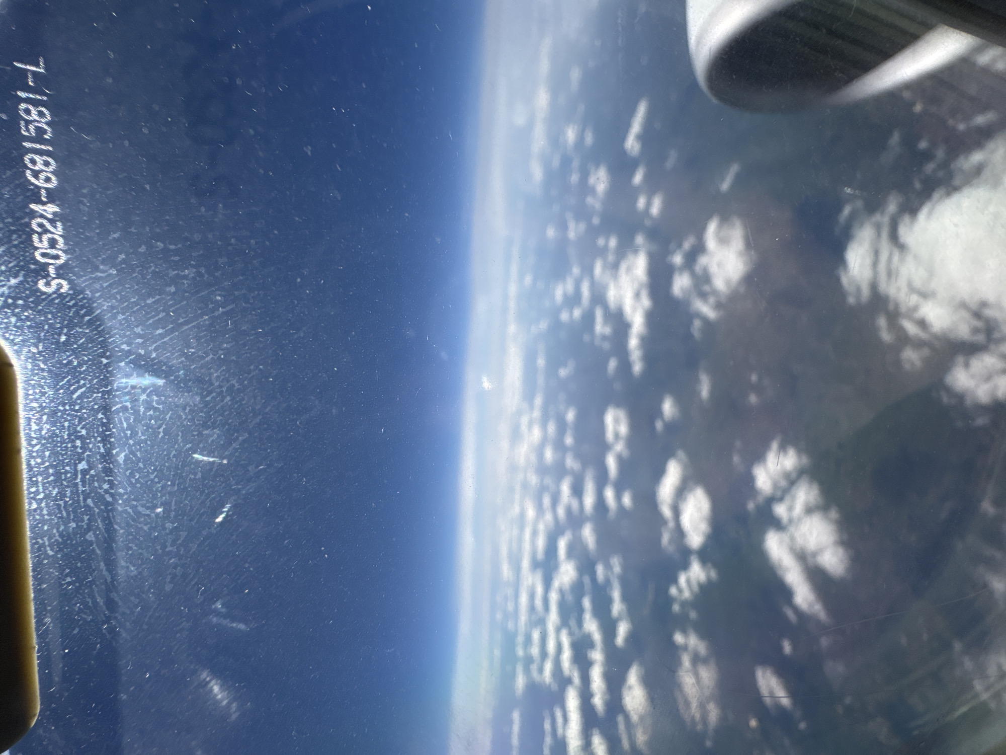 The image shows a view from an aircraft window, possibly during flight. The left side of the frame displays a blue sky transitioning to a lighter horizon. The right side shows a landscape of clouds. Part of the aircraft's wing or a structural element is visible in the top right corner. There is printed text "S-0524-681581-L" on the window. It is impossible to infer the city of Sofia, Bulgaria from the image.