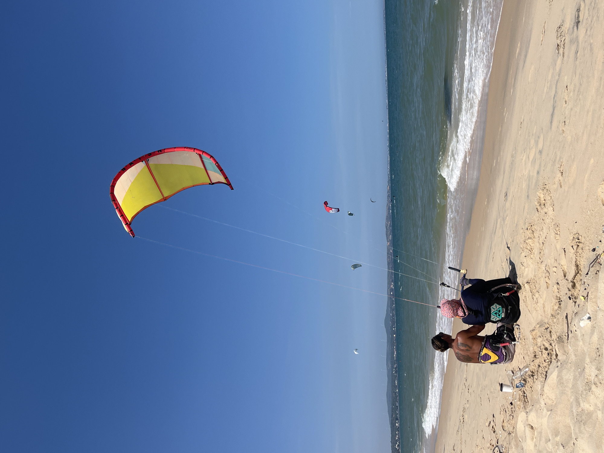 Two individuals are seated on a sandy beach, facing the ocean. The person on the left has their back to the camera, displaying a large tattoo on their back and wearing patterned shorts. The person on the right, who appears to be in a wheelchair, is wearing a pink headscarf and dark clothing. Both individuals are holding onto the controls for a kite. A large, multi-colored kite, predominantly yellow with red and white accents, is soaring in the clear blue sky, tethered by lines to the individuals. Several other kites are visible in the distance over the ocean, along with a coastline featuring buildings and wind turbines. The sea is a deep blue-green with moderate waves breaking on the shore. The beach is sandy and appears somewhat littered with various debris, including plastic bottles and wrappers. The scene suggests a location in Phu Thuy Ward, Vietnam, known for kite surfing activities.