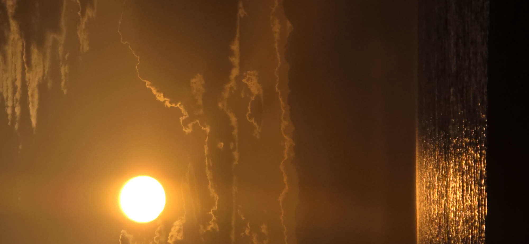 This is a vertical, full-shot photograph of a sunset over the sea in Boz Beshik, Kyrgyzstan. The sun, a bright white orb, is positioned in the upper left quadrant, casting a warm golden glow across the sky and the rippling water. Wispy clouds, illuminated by the setting sun, dot the sky. The lower portion of the image features a dark silhouette of land, likely a beach or coastline, bordering the shimmering sea.