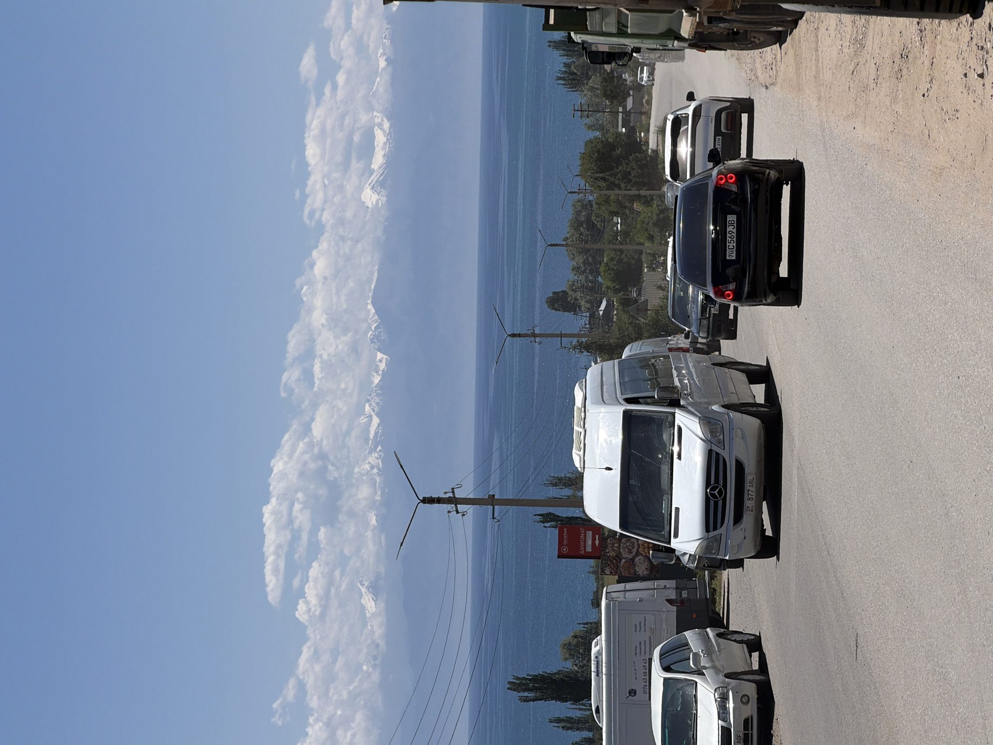 This media file captures a scenic view of a body of water, likely a lake, with snow-capped mountains in the background under a clear blue sky with scattered clouds. The image appears to be taken from a road alongside the water, which is lined with trees and some buildings.
Several vehicles are visible on the road, suggesting a traffic scene. These include a black sedan with license plate "70 C 569 JB", a white van with a Mercedes-Benz logo and license plate "05 877 ABL", and other white vans and cars further down the road. There are also utility poles with electrical wires. A sign with Cyrillic text that includes "BANKOMAT" is visible on the right side of the road.
The lighting suggests it is daytime, with bright sunlight casting shadows. The overall mood is serene, with the natural beauty of the landscape contrasting with the presence of vehicles and infrastructure. The image is oriented vertically, as if taken with a phone held upright.