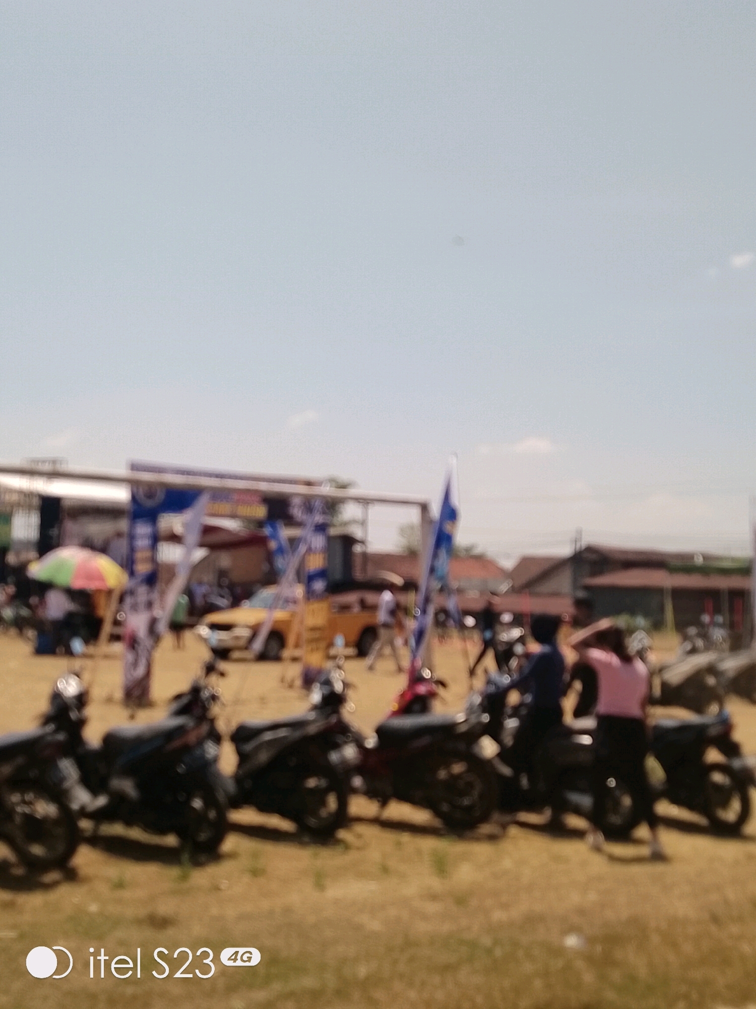 The image captures an outdoor scene in Blado, Indonesia, likely during a daytime event. A blue and white archway, possibly marking an entrance or stage, is visible in the background, adorned with banners. Several motorcycles are parked in the foreground, indicating transportation or a gathering. A yellow car is also present, suggesting a festive or community event. People are seen in the mid-ground and background, some interacting with each other and others appearing to be spectators. A colorful umbrella provides shade for a person on the left. The ground is covered in dry soil and patches of grass, typical of a rural or semi-urban environment in the region. The sky is clear and bright, suggesting fair weather. The overall impression is of a casual outdoor assembly or event.