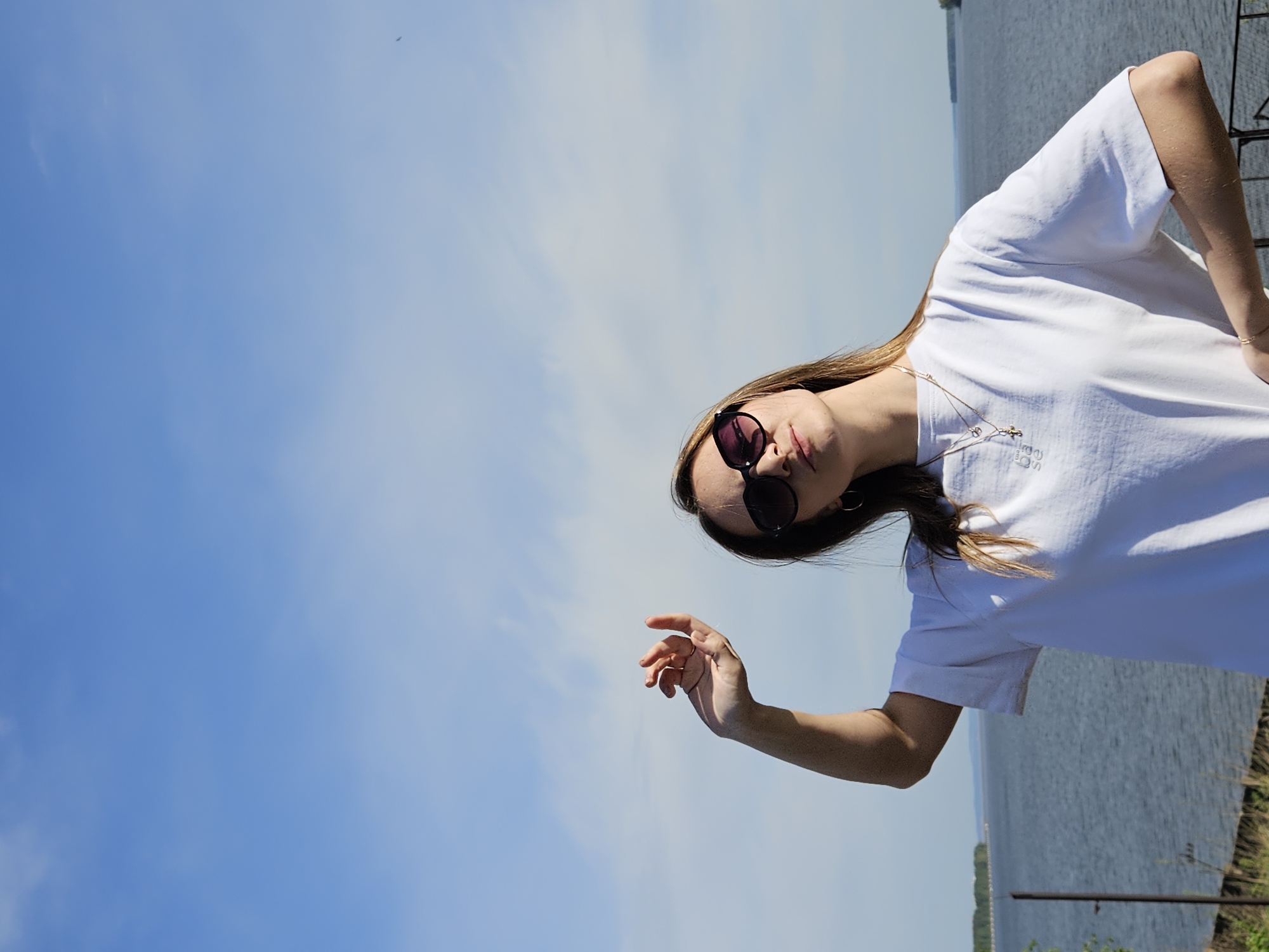 A woman in a white t-shirt and sunglasses stands outdoors, posing with her arm outstretched. In the background is a body of water, likely a river or large lake. The sky is blue with minimal clouds. Based on the location of Chaikovsky, Russia, the water may be part of the Kama River or a reservoir connected to it.