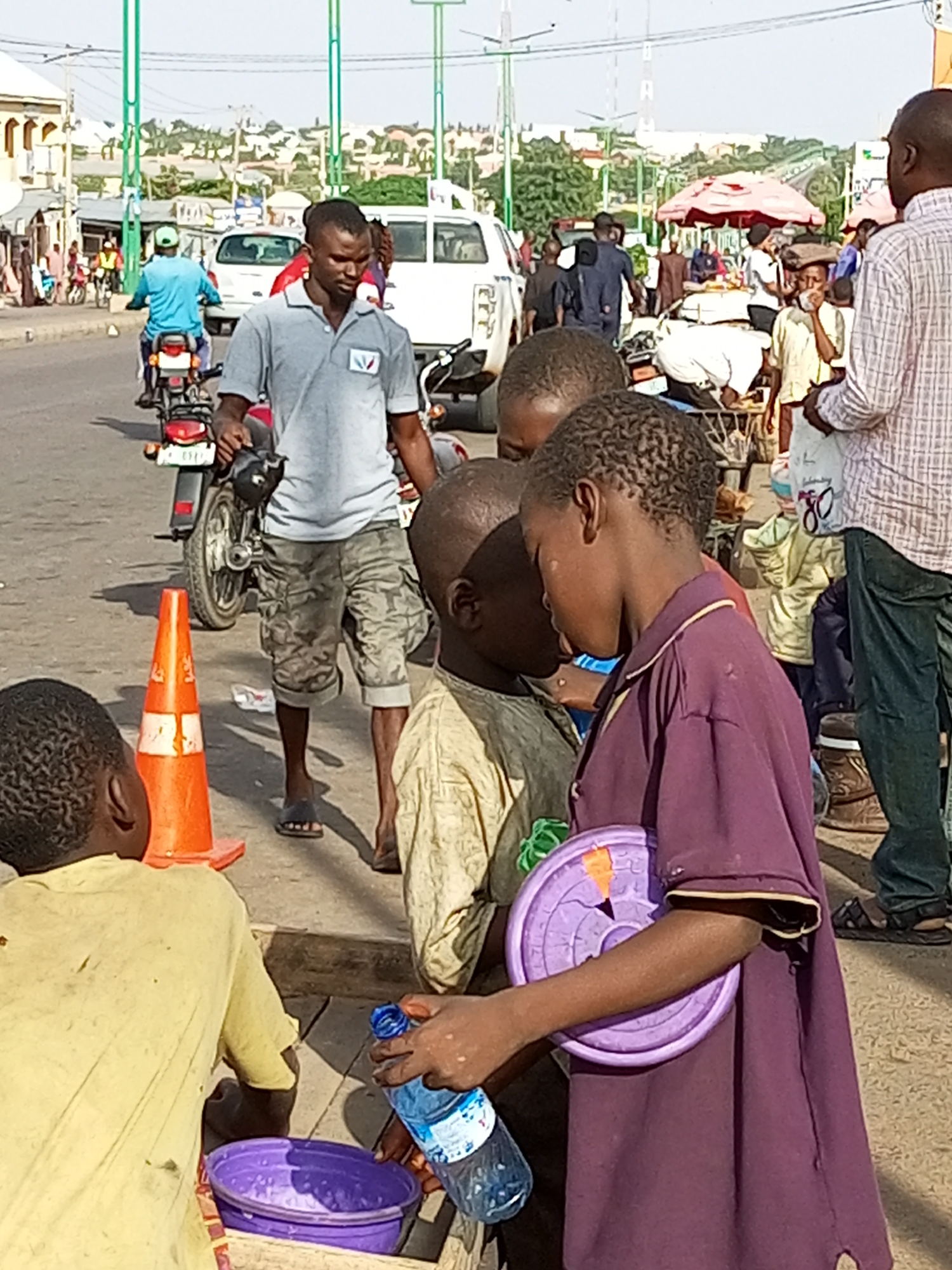 The image depicts a street scene in Tempure, Nigeria, characterized by a bustling urban environment with various individuals and vehicles. In the foreground, three young boys are gathered around a purple plastic bucket. One boy, wearing a yellow shirt, is partially visible from the back, interacting with the bucket. Another boy, in a light-colored garment, is looking down towards the bucket. A third boy, in a purple shirt, holds a purple plastic circular object and a partially full plastic water bottle.
Behind the boys, a man in a gray polo shirt and camouflage shorts stands near a motorcycle, holding a helmet. To his left, an orange traffic cone is positioned on the ground. Further in the background, a white van and other vehicles are present, along with a variety of people walking and standing. Some individuals are under a red umbrella, likely at a market stall. Buildings and trees line the street, suggesting a lively commercial or residential area. The overall atmosphere appears to be that of a busy public space on a sunny day.
