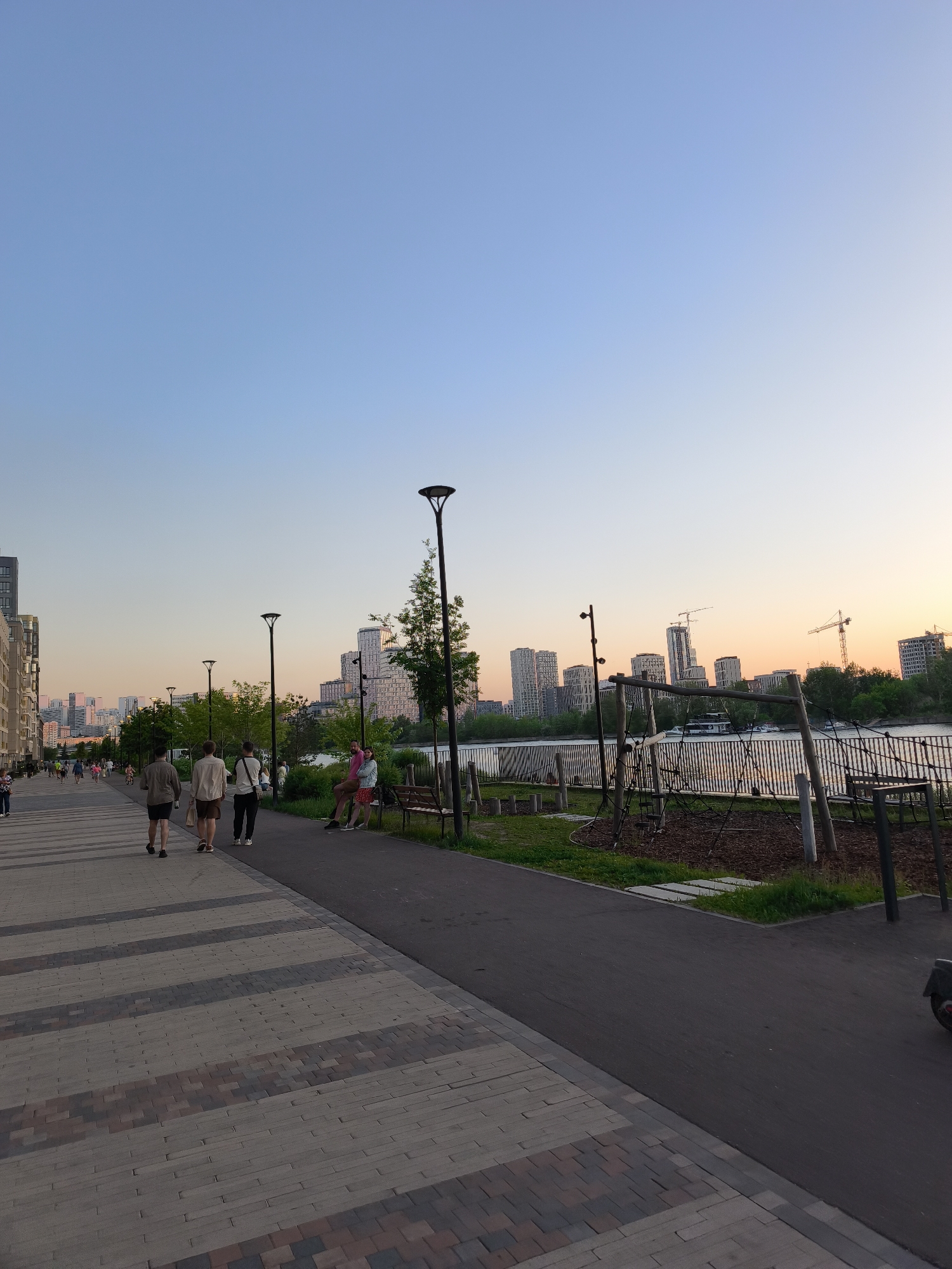 This image captures a wide, modern waterfront promenade in Moscow, Russia, specifically along the Karamyshevskaya Embankment, looking across the Moskva River towards the Shelepikhinskaya Embankment and its developing urban landscape. The scene is illuminated by the soft light of a late evening sunset, casting a clear sky that graduates from light blue overhead to pale orange and pink hues near the horizon on the right.
In the foreground, a broad pedestrian promenade on the left is paved with rectangular interlocking pavers in alternating patterns of light grey and darker grey-brown stripes, extending into the distance. Parallel to this, on the right, is a narrower asphalt path. Separating these two paths from the Moskva River is a strip of grass dotted with slender trees and evenly spaced modern, black streetlights featuring distinctive bell-shaped luminaires. Adjacent to the asphalt path on the right, a children's play area with wooden structures, ropes, and a wood-chip base is visible, bordered by a low metal fence running along the riverbank.
Several people are present. On the main promenade, three adult males are walking away from the viewer; the closest wears a dark t-shirt and shorts, the middle one a light shirt and shorts carrying a light bag, and the one on the right a dark top and trousers with a backpack. Further down the promenade, numerous other individuals are visible, walking and strolling. On the right side, two individuals are seated on a wooden slatted bench; one wears a pink top and shorts, and the other, partially obscured, wears a light jacket and a patterned dress. A third person stands near the bench. A fragment of a black wheeled scooter is visible in the extreme bottom right corner.
Across the Moskva River, the background is dominated by a dense array of modern, high-rise residential buildings, characterized by light-colored facades and distinct architectural designs, including some with curved or cylindrical elements. Several prominent construction cranes are interspersed among these buildings, indicating ongoing development. A white boat is seen on the river, while a line of green trees fringes the far bank beneath the distant buildings.