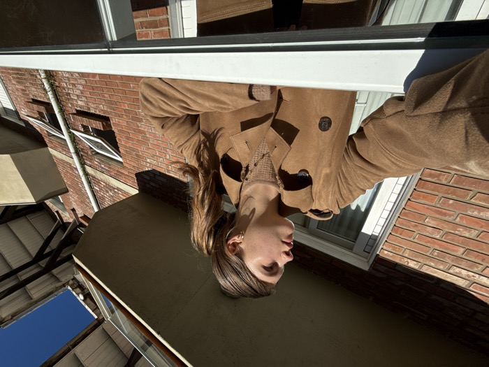 A low-angle, outdoor image depicts a young individual, appearing female, with light brown hair in a ponytail, looking upward and to the right. She wears a tan trench coat over a textured brown sweater. Her right arm extends forward, with a hand visible near the left edge of the frame, appearing to hold a device for photography. She is leaning over a prominent white horizontal railing or window frame in the foreground. Behind her, a multi-story building constructed of red bricks is visible. Two windows with white frames are open, tilted outwards from the bottom. A white vertical downspout runs along the brick wall. Above the subject, a concrete balcony structure projects outwards, casting a shadow. Further right, a section of a building with a light-colored roof supported by exposed wooden beams is present. A clear, bright blue sky is visible in the upper right portion of the frame. The scene is located in the city of Canela, Brazil.