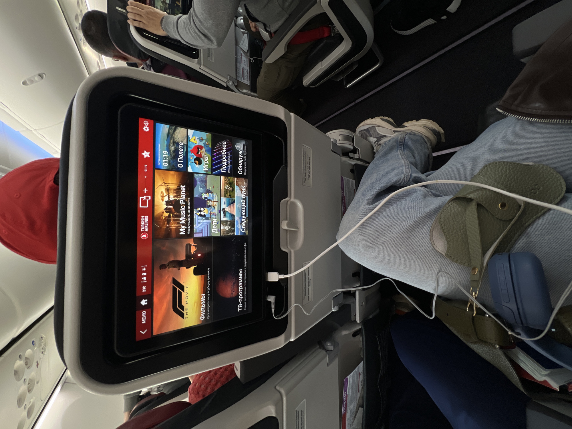 The image provides a first-person perspective from an airplane seat, focusing on an in-flight entertainment (IFE) screen. The screen displays a menu primarily in Russian, featuring options such as "О Полете" (About the Flight), "Игры" (Games), "Фильмы" (Movies), "ТВ-программы" (TV Programs), "My Music Planet" (Моя музыкальная планета), and sections for "Дети" (Children) and "Подробнее" (More details). The "TURKISH AIRLINES" logo is prominent at the top of the screen, and a "F1 THE MOVIE" tile is visible. The remaining flight time is displayed as "01:19".
A white charging cable is plugged into the IFE system, extending towards the lap of the person taking the photo, who is wearing light blue jeans and grey and white athletic shoes. On their lap, an olive green textured pouch with a gold clasp and a blue hard case with an attached white cable are visible.
In the seat directly ahead, another passenger is seen from behind, wearing a grey long-sleeve shirt and dark pants, with their hands resting on the seat in front. A red seatbelt strap is also visible. Partially obscured to the left of the screen is another passenger wearing a red cap. Overhead panels with cabin lights and air vents are visible in the background. The setting is an airplane cabin during a flight. Text on a partially visible sticker on the seat structure includes "757-9 MAX," likely indicating the aircraft model. The scene depicts a calm and routine in-flight experience.