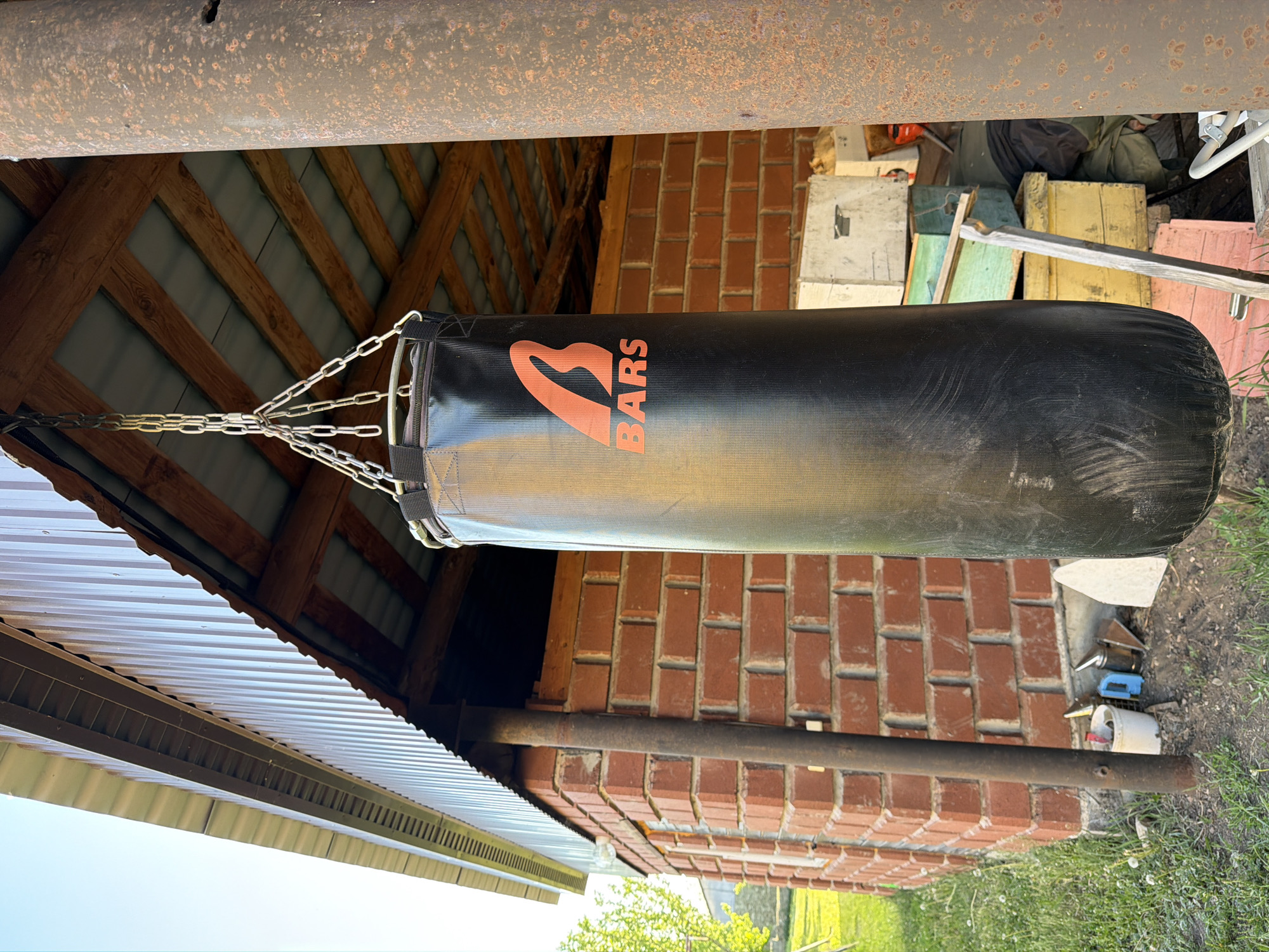 A cylindrical black punching bag with an orange logo is suspended by chains from a rusted metal beam under a wooden-framed structure. The wooden structure is partially covered by corrugated metal roofing on one side, and a brick wall is visible on another. Adjacent to the brick wall are miscellaneous items including scrap wood and various containers. The surrounding environment includes grass and vegetation. The location is Bolsheustyikinskoye, Russia.