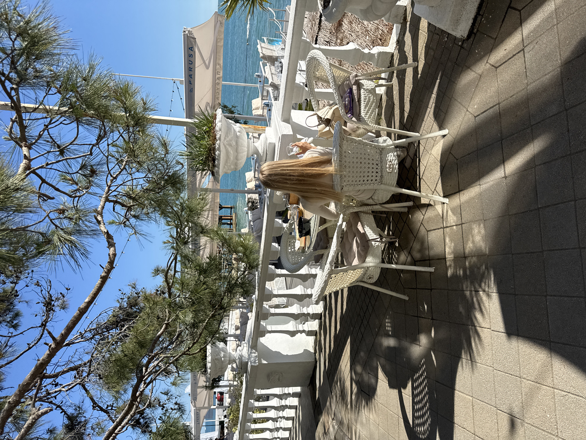 An outdoor scene captures a sunny terrace cafe or restaurant by the sea, in Sochi, Russia. The image is taken on a bright, clear day with a vibrant blue sky. In the mid-ground, a person with long, straight blonde hair is seated at a white wicker table, viewed from behind. They are holding a clear glass, seemingly enjoying a drink or a meal. An adjacent white wicker chair holds a light-colored bag.
The terrace features a distinctive white balustrade and decorative white planters filled with green plants. Beyond the balustrade, the calm, blue waters of the sea are visible, confirming a coastal setting. Several white awnings or parasols provide shade further back. Overhead and to the left, the branches of a pine-like tree with long needles are prominent. The strong sunlight casts long, distinct shadows of the chairs, balustrade, and trees onto the tiled patio floor, indicating either morning or late afternoon.
Visible text on one of the awnings reads "PARUSA", with what appears to be reversed text "ACYRAG" nearby. The overall ambiance is peaceful and relaxing, typical of a sunny day at a seaside resort.