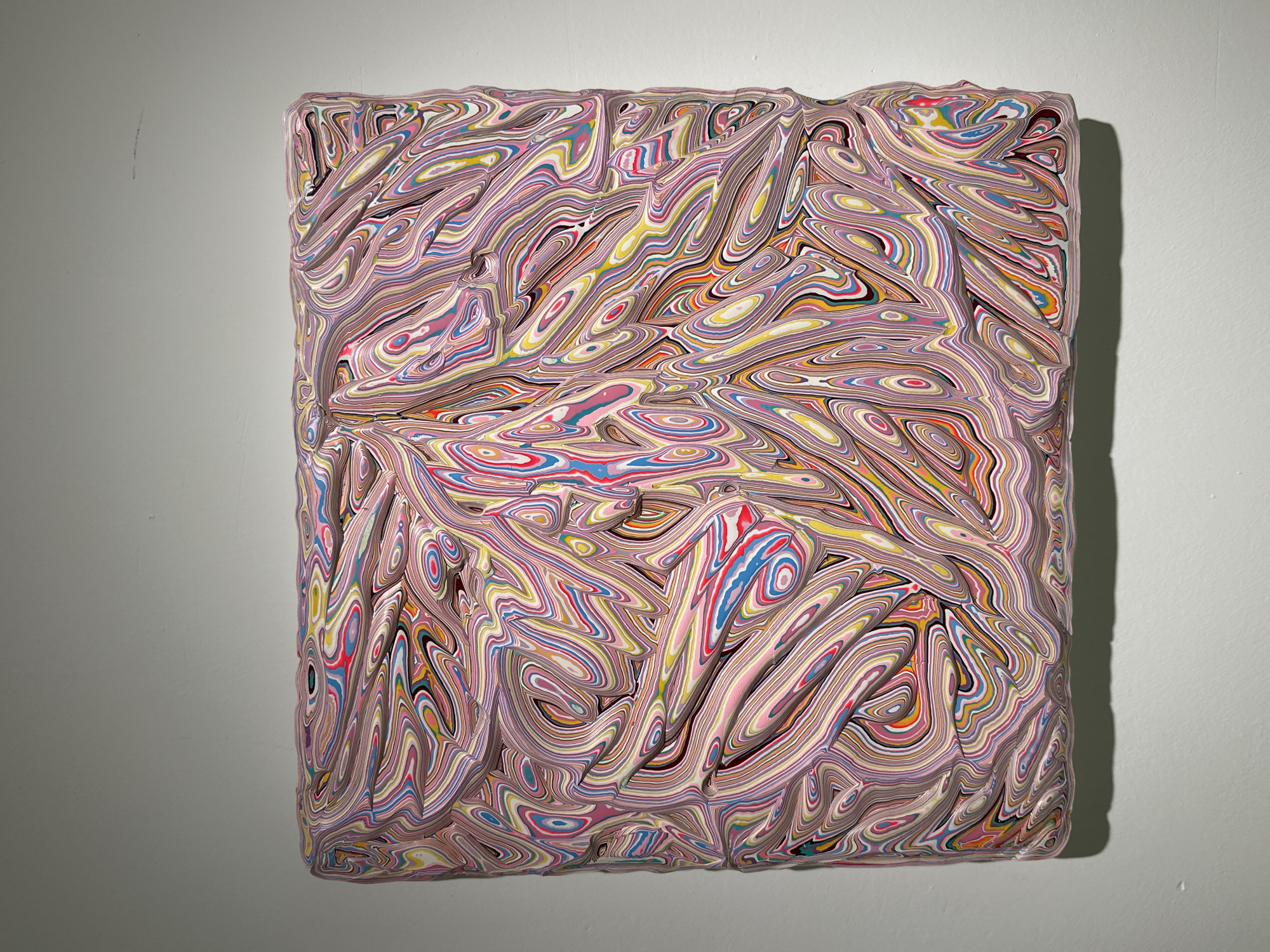 A square abstract artwork hangs on a white wall. The artwork is composed of layers of swirling colors in a pale pink base, including white, yellow, red, and blue. The layered colors create a three-dimensional effect with undulating ridges and valleys. The artwork is positioned against the wall, casting a shadow on the right side. The wall appears smooth and unmarked except for the hanging artwork. The location is not discernible and unrelated to Banan, China.