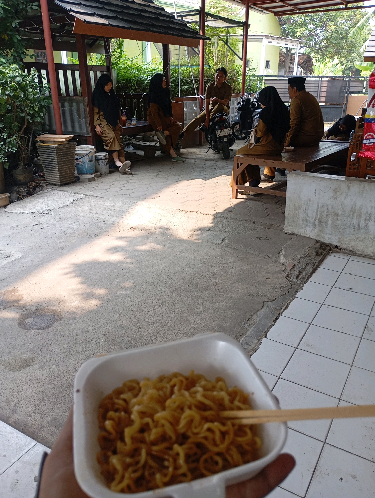 In Babelan, Indonesia, a casual outdoor gathering takes place under a covered pavilion. A person is holding a white bowl filled with instant noodles, with wooden chopsticks resting on the rim. In the background, several individuals are seated on a wooden bench. Two women wearing hijabs and long brown dresses are seated on the far left of the bench. Further to the right, a man in a brown uniform is sitting on a motorcycle. Next to him, a man and a woman in brown uniforms are seated on the bench. Behind the people, a motorcycle is parked. The ground is a mix of concrete and interlocking pavers. Sunlight casts shadows on the ground. In the distance, a building and trees are visible.