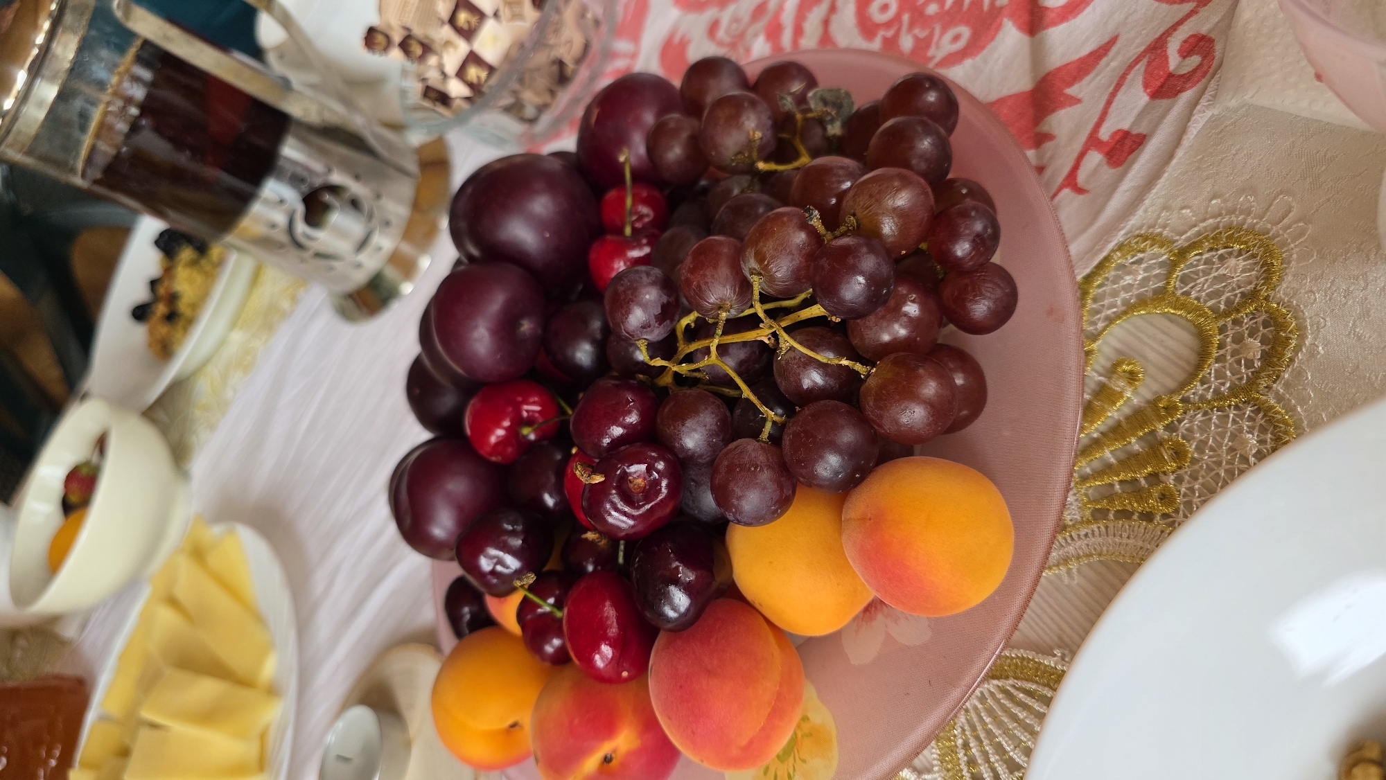 A close-up, eye-level shot captures a platter of fresh fruit on a table covered with a pink and white patterned tablecloth. The platter, made of pink glass, is filled with a vibrant assortment of red grapes, dark red cherries, and orange apricots. The fruits are arranged in a natural, appealing way, suggesting a leisurely meal or snack. In the background, slightly out of focus, are other elements of a meal setting. To the left, a white plate holds sliced yellow cheese. Further back, a white bowl contains a mix of fruits, and a clear glass French press with dark liquid stands to the right. The scene is brightly lit, possibly by natural light, highlighting the textures and colors of the fruits. The overall impression is one of abundance and a healthy, fresh offering. The exact location in the city of Купоярово, Russia, cannot be determined from the visual information provided.