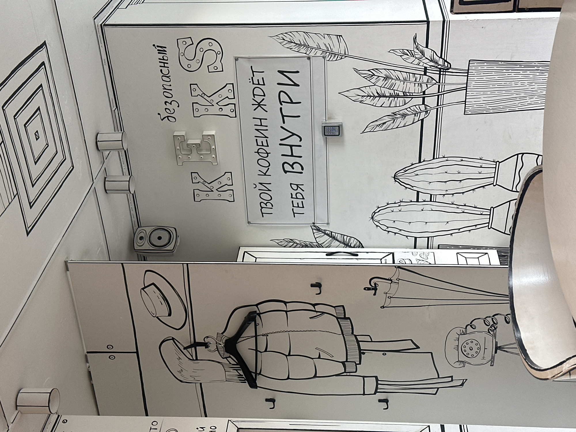 The image shows the interior of a cafe with a hand-drawn, cartoonish aesthetic. The walls and ceiling are decorated with black outlines, creating a 2D effect. In the foreground, a portion of a light-colored chair is visible. On the wall, there is a drawing of a jacket hanging on a hanger, a hat, and a rotary telephone. Further to the right, there are drawings of potted plants, including cacti and large-leafed plants. A sign in Russian reads "ТВОЙ КОФЕИН ЖДЁТ ТЕБЯ ВНУТРИ" which translates to "Your caffeine is waiting for you inside". The cafe is located in Minsk, Belarus.