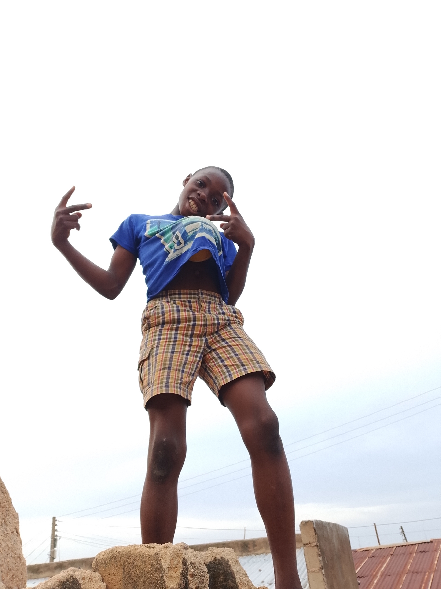 A low-angle shot captures a young boy, likely under ten years old, standing on a pile of concrete blocks. He wears a blue t-shirt and plaid shorts, with his arms outstretched and hands held up in a "rock on" gesture. The boy grins widely, revealing his teeth. The background is a plain, overcast sky, suggesting an outdoor setting in an urban area. In the distance, power lines and a corrugated metal roof are visible, hinting at a developing or industrial environment. The boy's knees show some scuff marks, indicating recent activity. The overall impression is one of youthful exuberance and confidence against a simple, possibly impoverished backdrop.