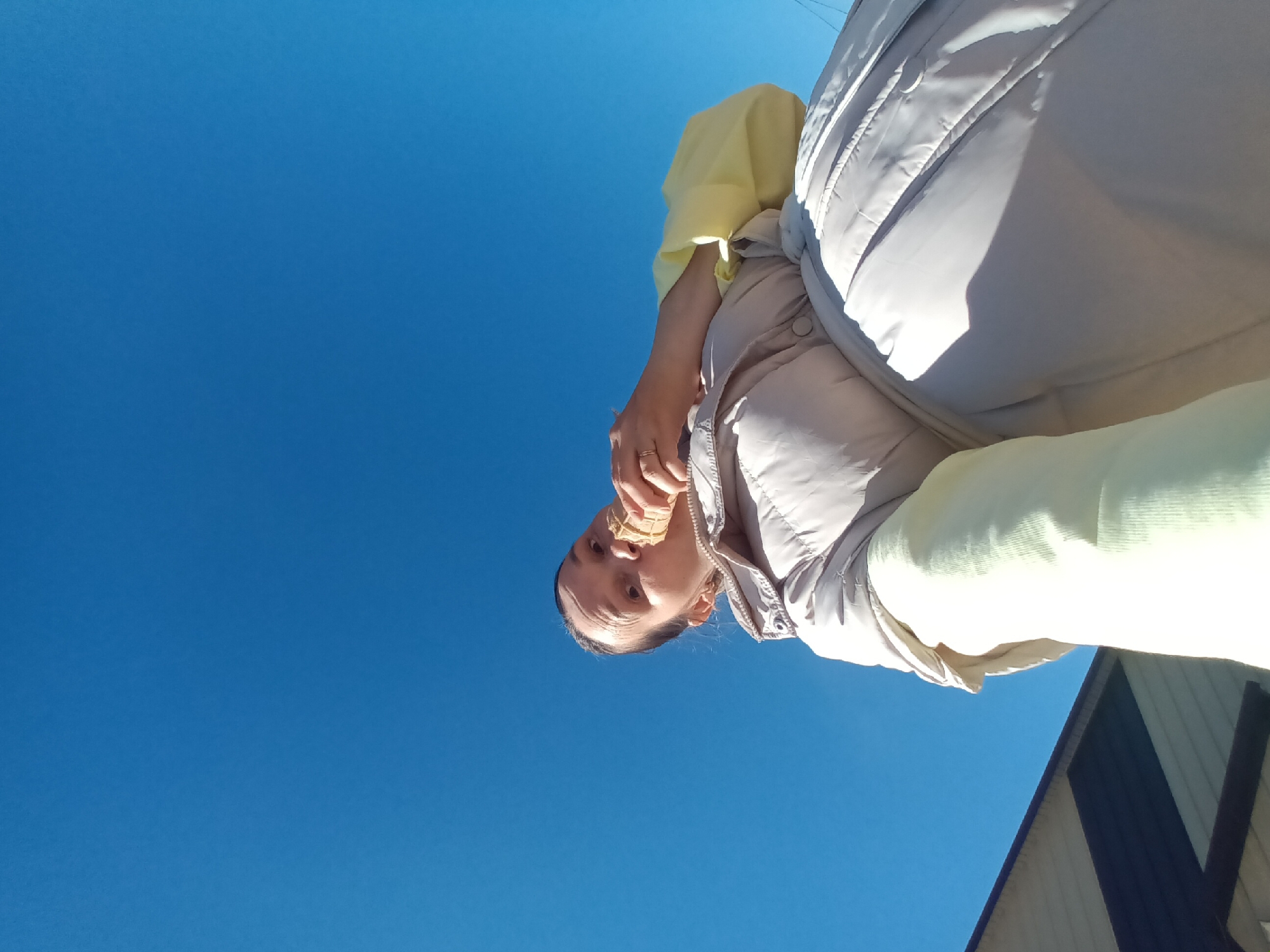 A low-angle shot depicts a person eating an ice cream cone against a clear blue sky. The person is wearing a light yellow long-sleeved shirt and a light beige padded vest. They appear to be standing outdoors in Ольховка, Russia, though no specific landmarks confirm the location. The bottom right corner of the image shows a portion of a building or structure with a dark roof or siding, contrasting with the sky's uniformity. The focus is on the person and their action of eating, emphasizing the upward angle and the expansive blue backdrop.