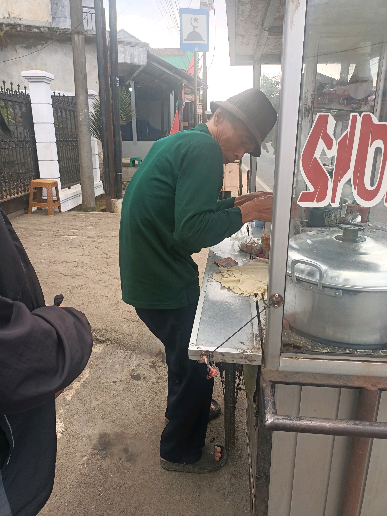 In Sukamukti, Indonesia, an elderly man wearing a green long-sleeved shirt and a brown hat is preparing food at a street cart. The cart features a large silver pot with a lid. Red lettering is visible on the glass display of the cart. The ground is paved with concrete, and a black jacket and a portion of black pants are visible in the foreground on the left. A small, brown stool sits near a fence on the left side of the image. Electrical poles and a building with a grey facade are in the background.