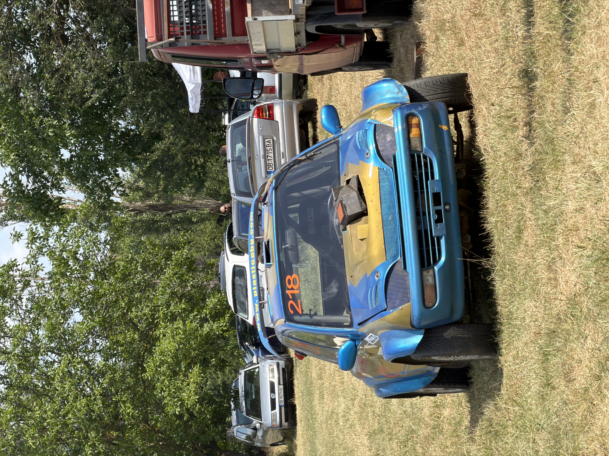 In Bahovitsa, Bulgaria, a modified blue and yellow rally car with the number 218 prominently displayed on its windshield is parked on a dry grassy field. The car has an exposed engine compartment with a black intake box. Other vehicles, including a silver Volkswagen Passat, a silver Opel Astra, and a red truck, are visible in the background. Several people are standing around the vehicles. The scene is set in an outdoor environment with trees and foliage surrounding the area. The sky is partially visible and appears to be clear.