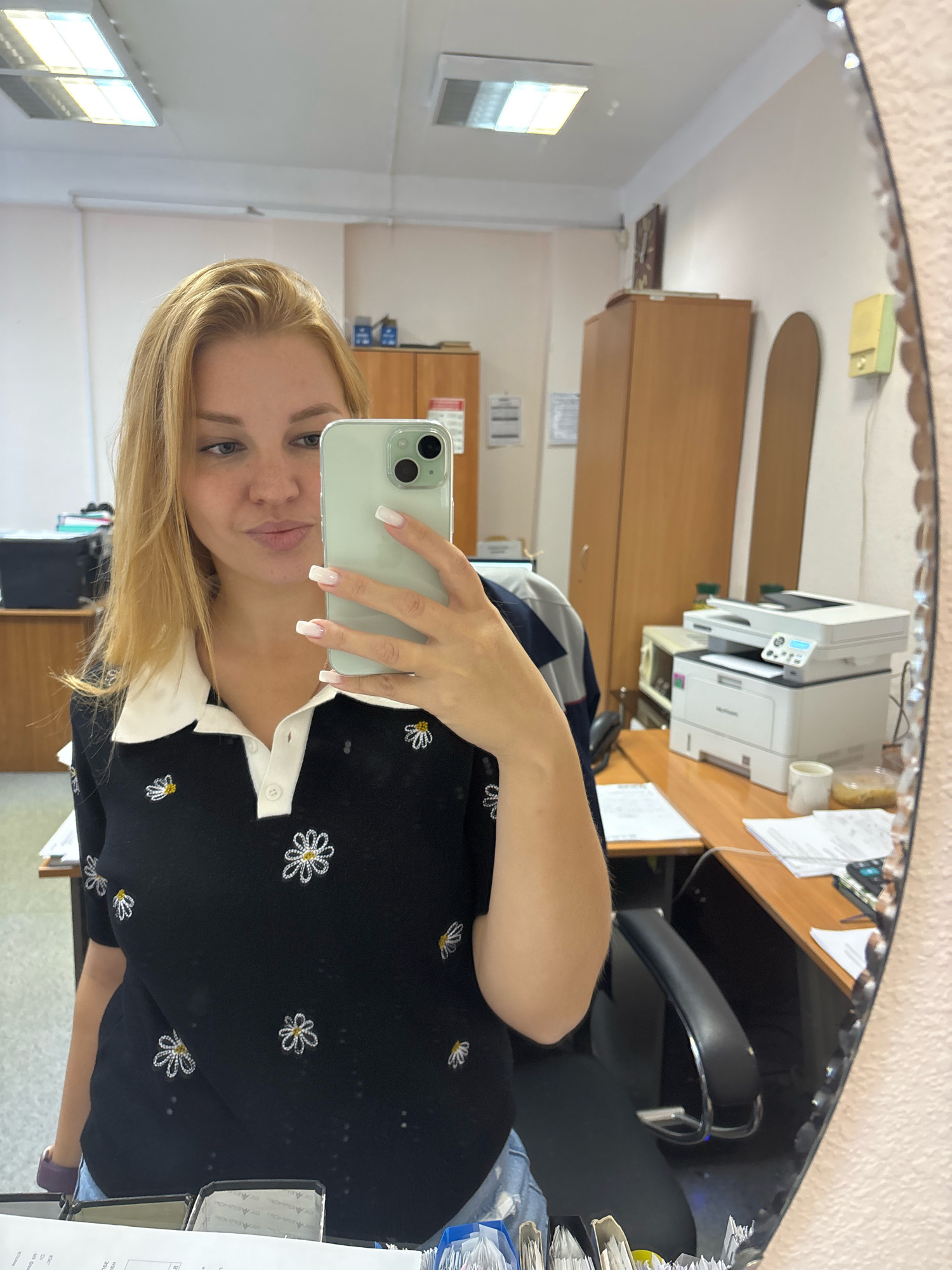 A young blonde woman with white-tipped fingernails takes a mirror selfie in what appears to be an office. She is wearing a black collared shirt with a white collar and a pattern of small embroidered white daisies. She is holding a light green smartphone in her right hand, and her left arm is visible. In the background, reflected in the mirror, are office furniture, including cabinets, desks, a printer, and office chairs. The environment suggests a workplace setting. The location is specified as Rusanovo, Russia.