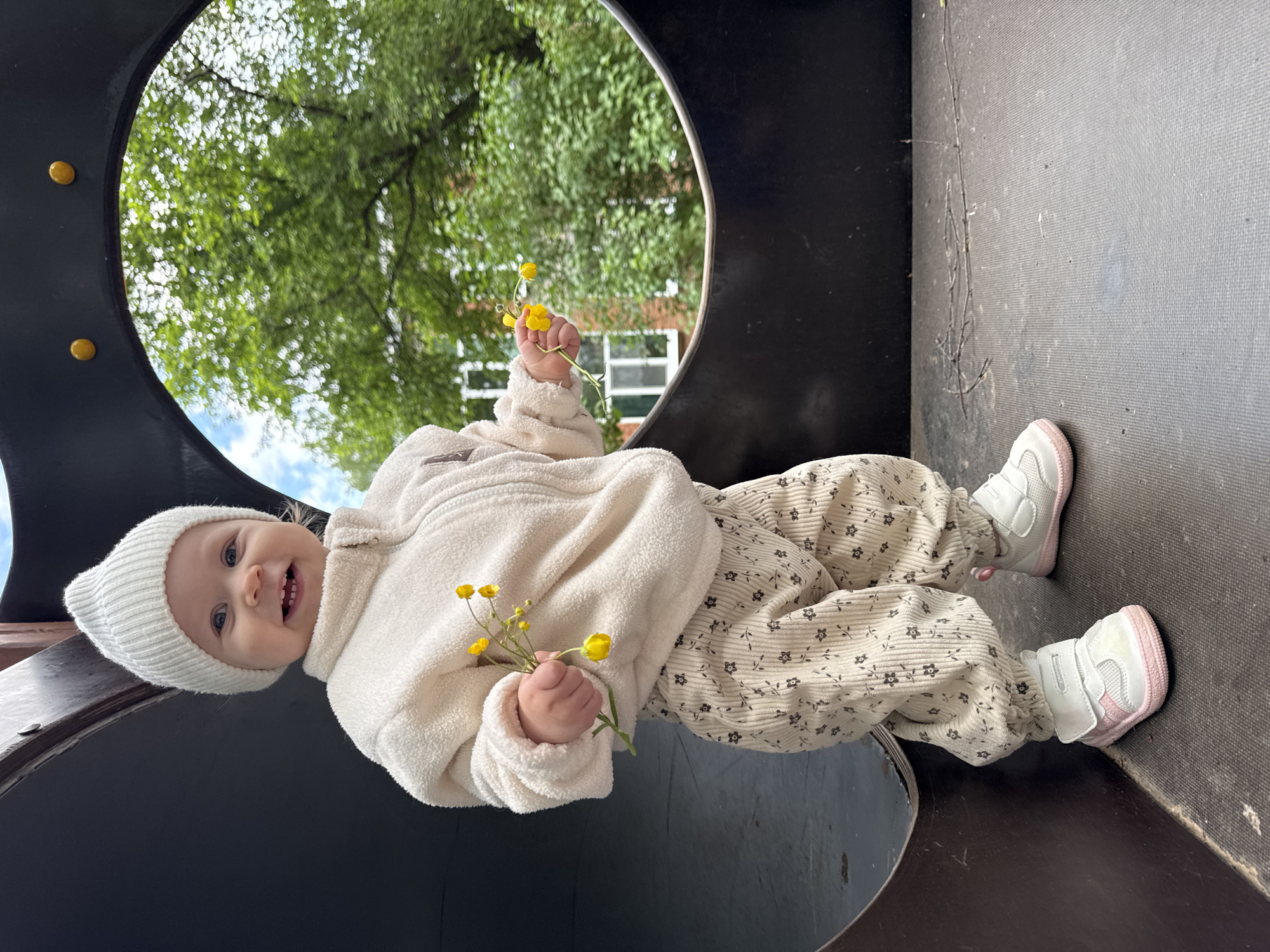 A baby wearing a white knit hat, a cream-colored jacket, and patterned corduroy pants, is smiling and holding yellow flowers. The baby is inside a play structure with a circular opening. Green trees and the sky are visible through the opening. The surface the baby is on appears to be dark gray. The location is not specified in the image.