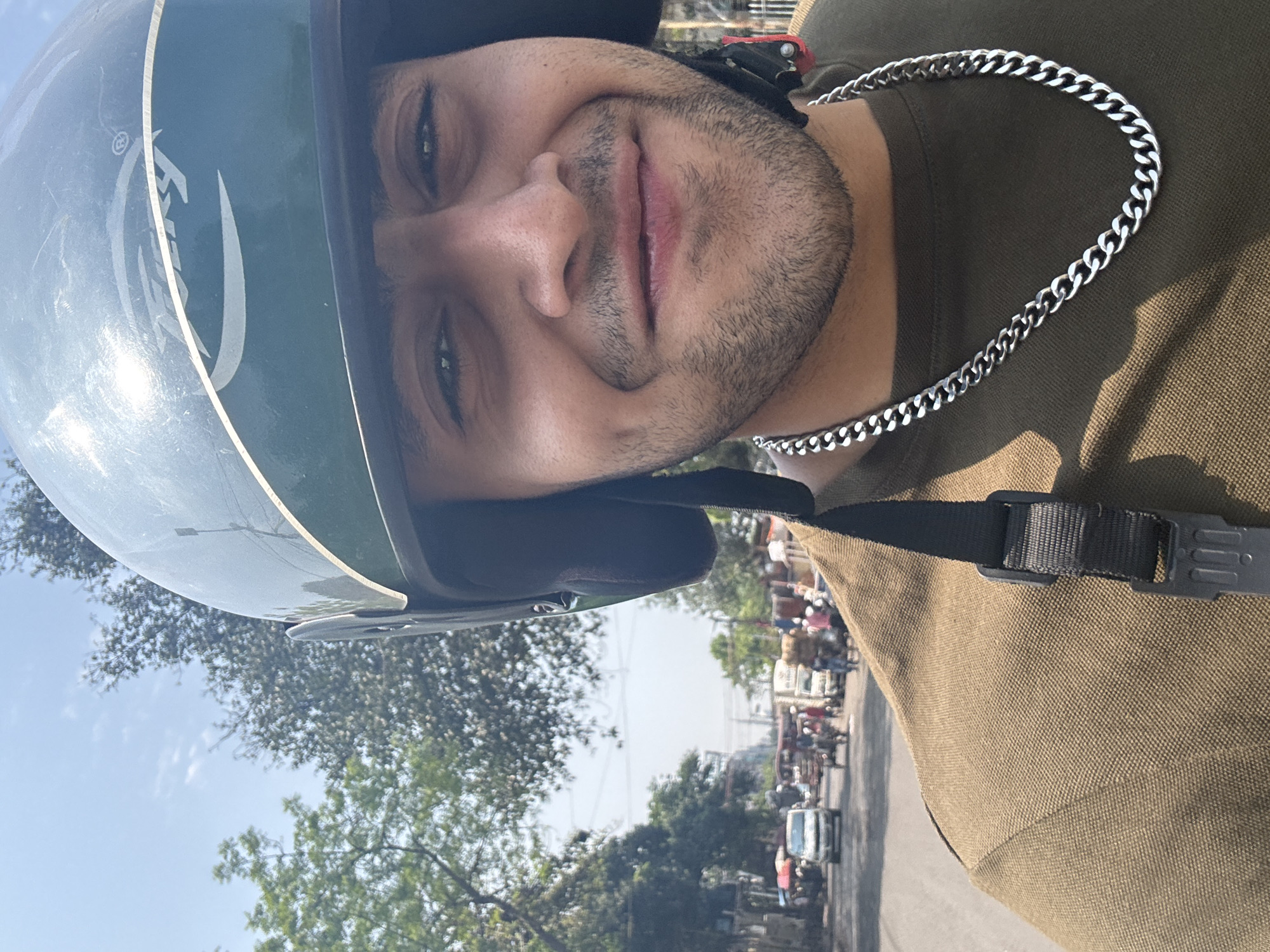 A close-up photograph features an adult male, likely in his 20s or 30s, wearing a dark green motorcycle helmet with a light-colored stripe and "TIEREY" branding on the side. His helmet's chin strap is fastened. He has short facial hair, dark eyes, and is smiling directly at the camera, exhibiting dimples. He is wearing an olive-green polo shirt and a silver-colored cuban link chain around his neck. The background depicts a busy urban street scene under a clear blue sky with scattered clouds and visible green foliage from trees. Various vehicles, including cars, motorcycles, and commercial vehicles (some laden with goods), are visible on the road, alongside numerous pedestrians. This scene is consistent with a bustling street in the city of Kolkata, India.