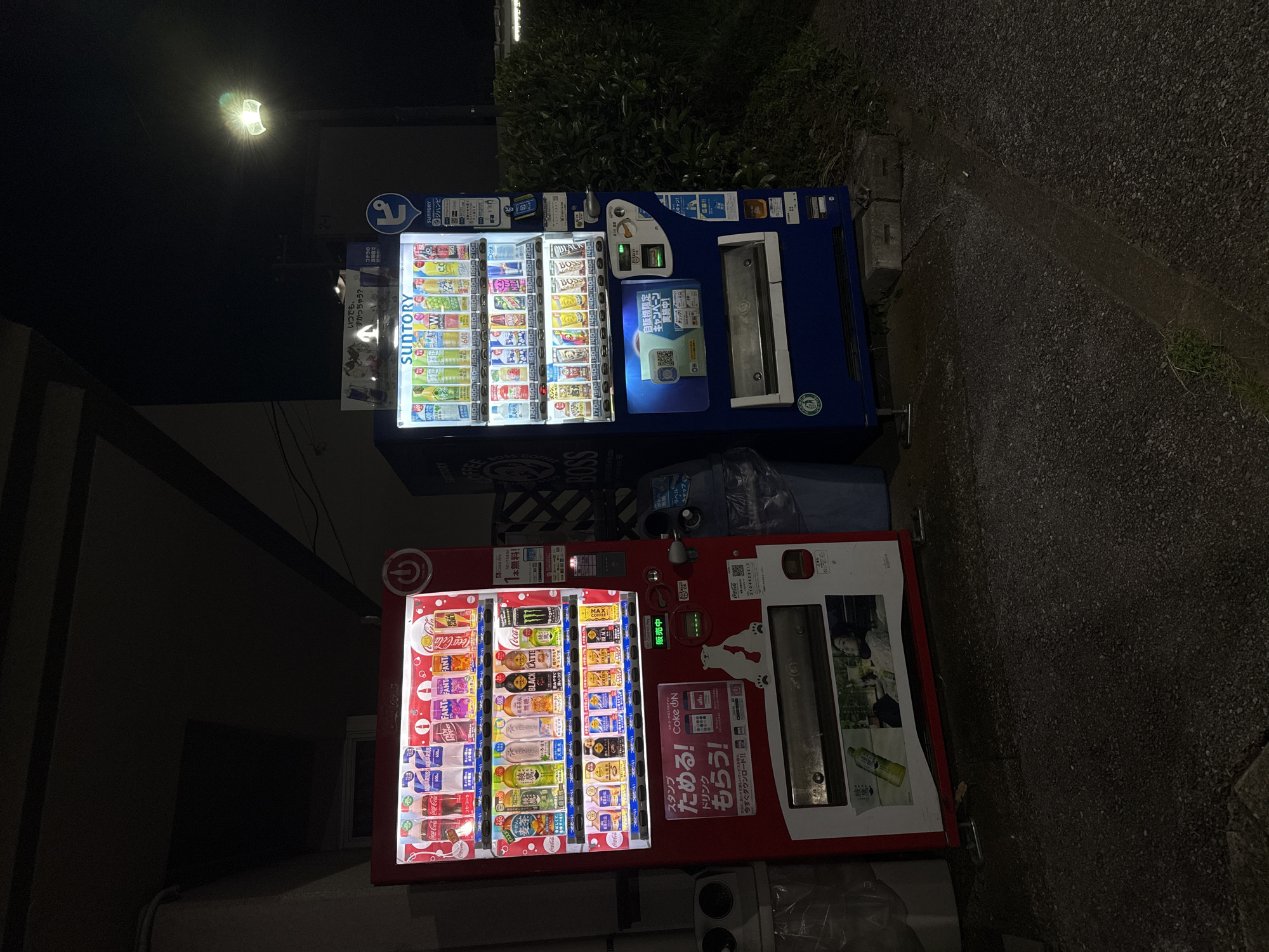 The image shows two vending machines at night in Yachiyo, Japan. The vending machine on the left is red and features Coca-Cola branding. It dispenses various drinks, including Coca-Cola, Aquarius, Dr. Pepper, and Fanta. There is also an advertisement for "Coke ON" and a graphic of a polar bear. The vending machine on the right is blue and branded with "SUNTORY" and "BOSS." It dispenses a variety of Suntory beverages, including teas and coffees. Both machines are illuminated by internal lighting. In the background, there is a street lamp and dense green foliage. The ground surface is asphalt with some gravel. A trash can is visible between the two vending machines.