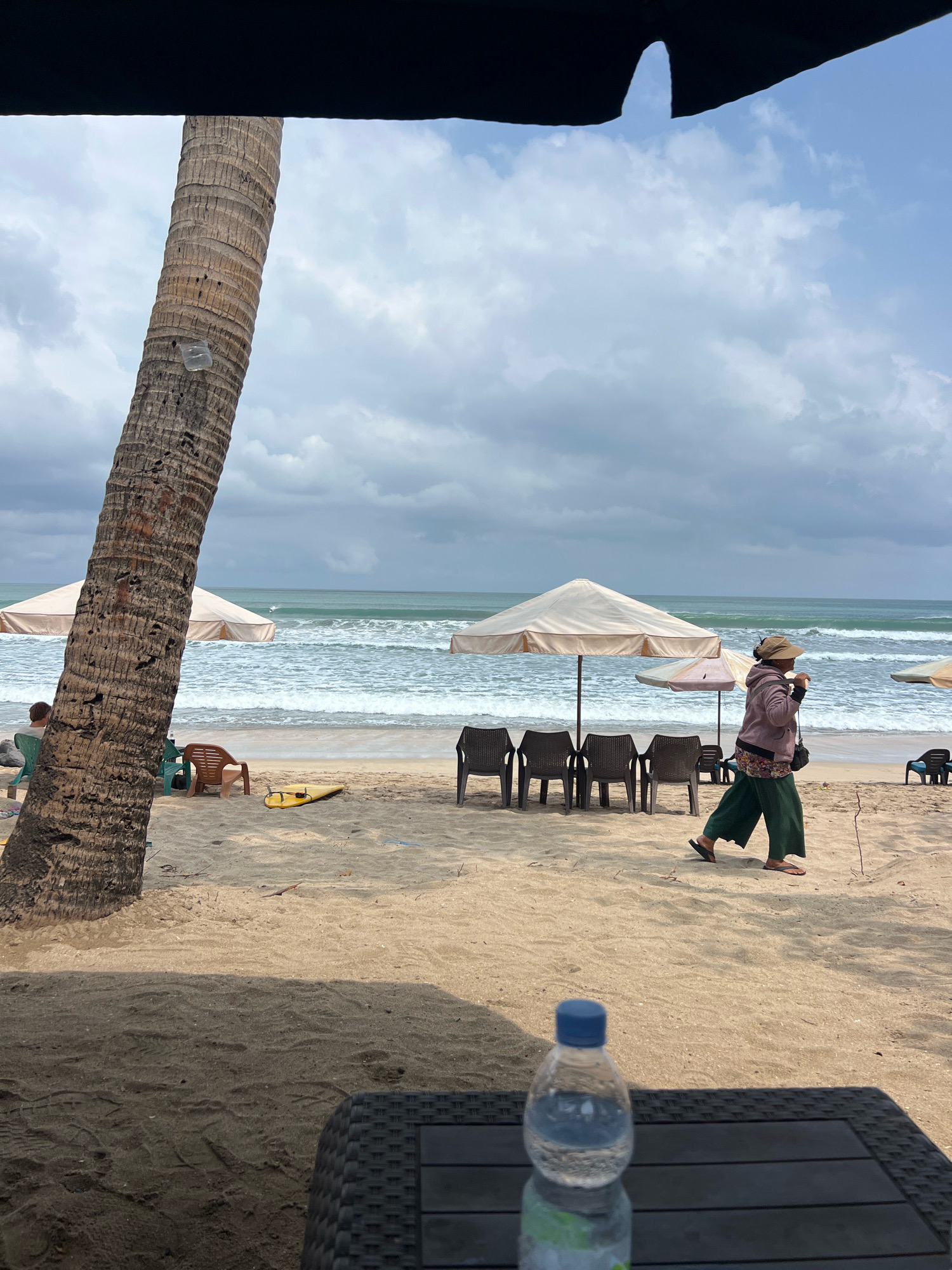 A person walks across a sandy beach in Kuta, Indonesia, under a cloudy sky. The individual, wearing a wide-brimmed hat, a pink hooded jacket, and loose green pants, is moving from right to left, away from the ocean. In the foreground, a plastic water bottle rests on a dark, woven surface, possibly a table.
The beach is lined with beach chairs and umbrellas. A large palm tree trunk dominates the left side of the frame, partially obscuring the view. Beyond the chairs, the ocean stretches out, with moderate waves rolling towards the shore. A yellow surfboard lies on the sand near some chairs. Further in the distance, a surfer is visible in the water. The overall scene suggests a tranquil beach setting, despite the overcast weather.