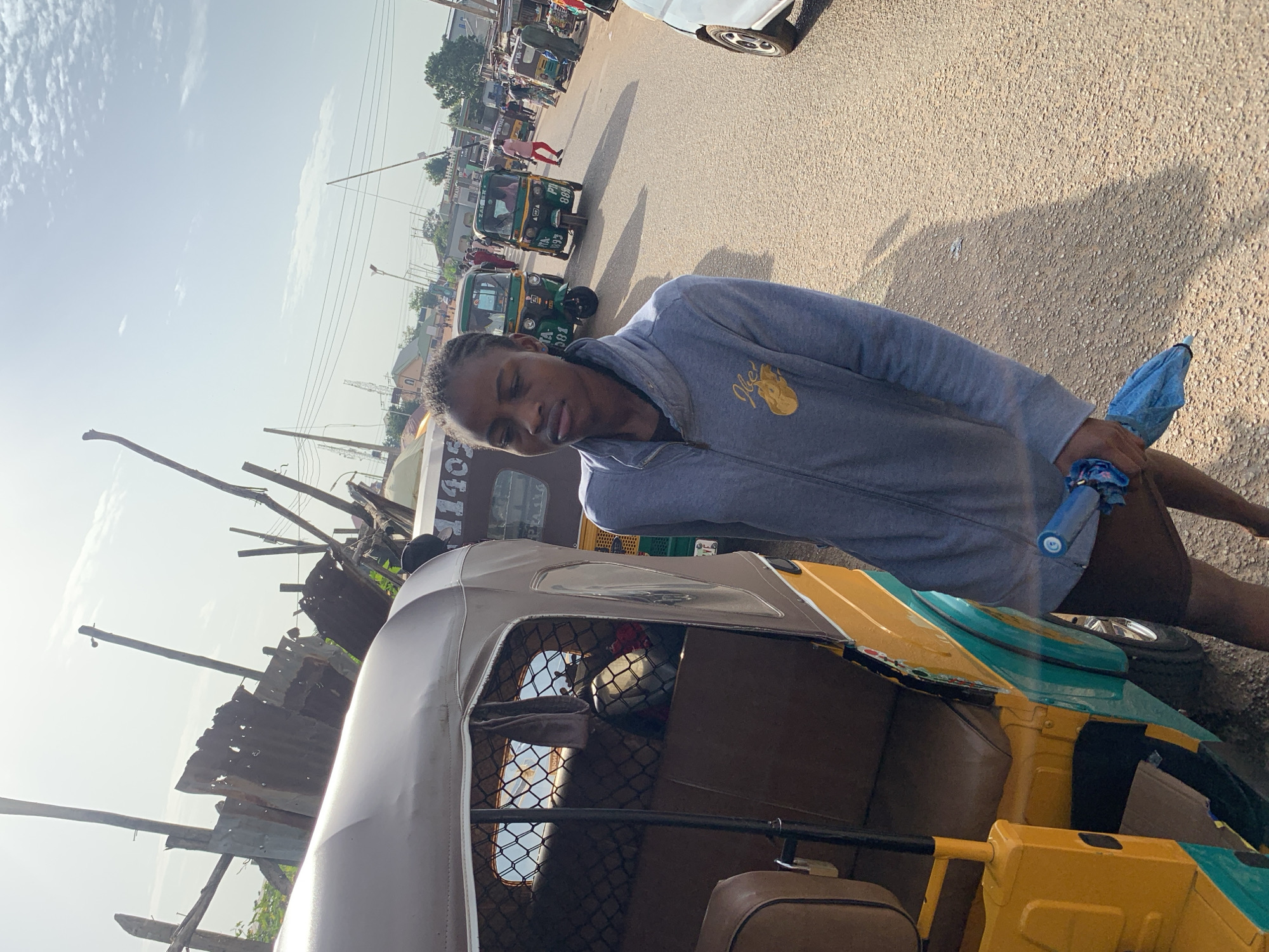 A young Black person stands next to a yellow and teal auto-rickshaw on a dusty street in Gura Top, Nigeria. The person, wearing a grey hooded jacket and dark shorts, holds a folded blue umbrella in their right hand. The auto-rickshaw has a brown canvas roof and brown seats. In the background, other auto-rickshaws are visible, along with pedestrians and simple structures made of corrugated metal and wood. The sky is bright and partly cloudy. The scene appears to be a busy street market or transportation hub.