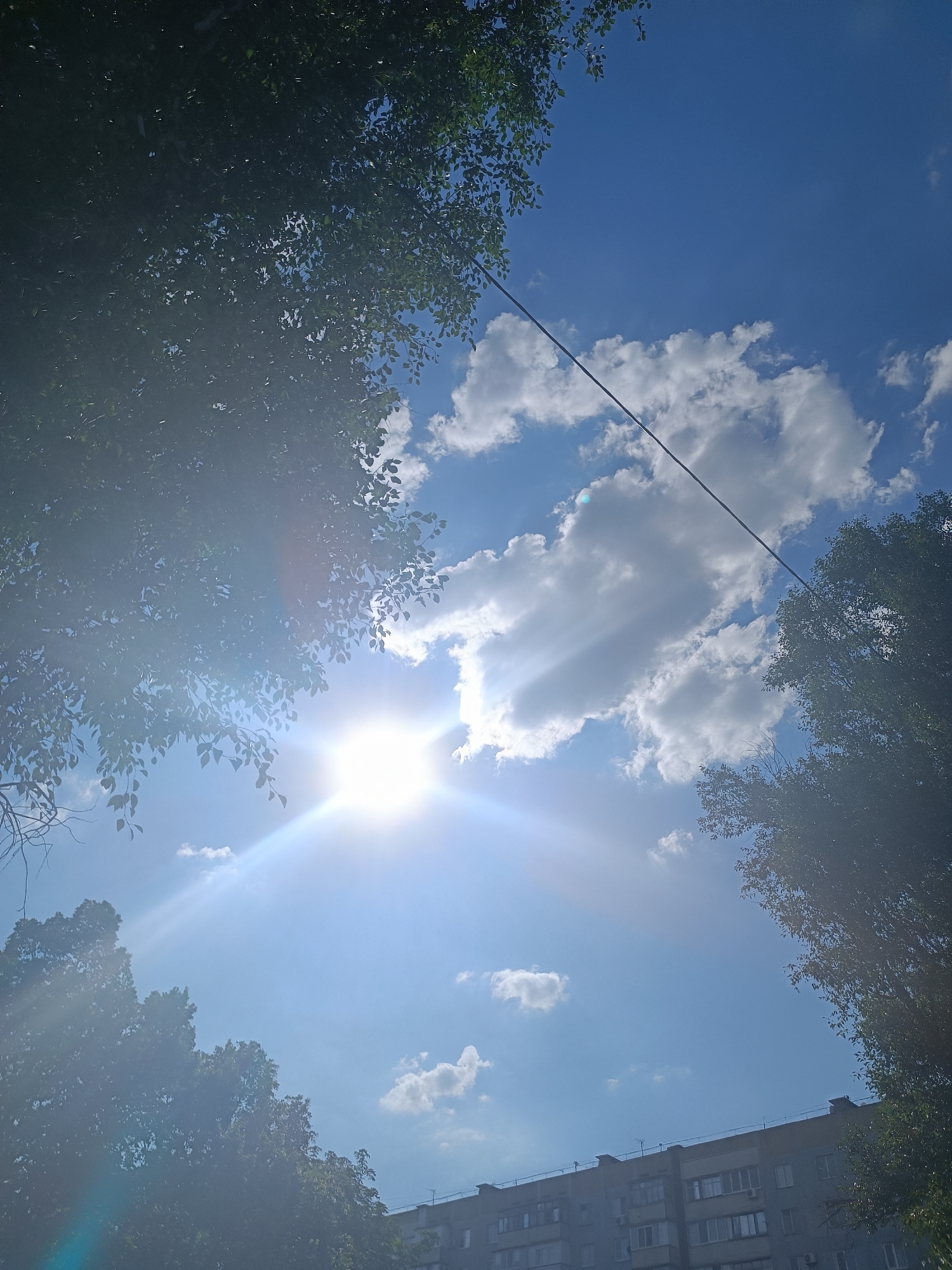 A clear blue sky is dotted with fluffy white clouds. The sun shines brightly, creating lens flare and casting rays of light through the atmosphere. Silhouettes of trees frame the scene from the top left and right, their leaves obscuring parts of the sky. A thin black wire stretches diagonally across the upper portion of the image. In the bottom right corner, the upper section of a grey, multi-story apartment building is visible, with visible windows and air conditioning units. The setting appears to be an urban environment on a sunny day.