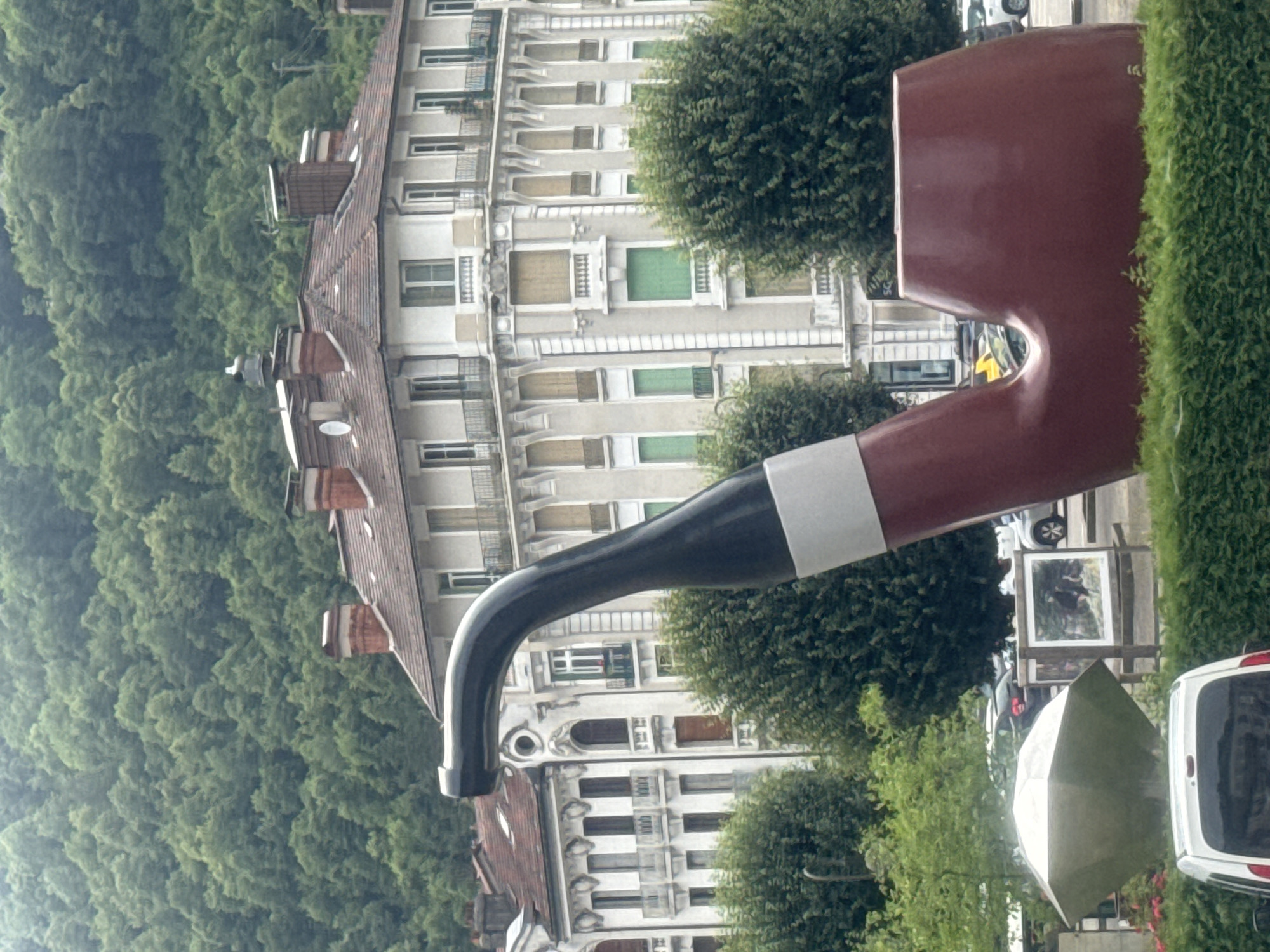 The image shows a large, reddish-brown sculpture of a pipe in the foreground. The pipe sculpture has a black stem with a white band. Behind the pipe, several multi-story buildings with ornate architecture and pitched roofs are visible. Trees and a forested hillside form the background. In the lower part of the image, a white vehicle, a polygonal-shaped sculpture, and a framed picture are situated behind a hedge. The scene appears to be in an urban or town setting, possibly a park or public square, given the sculptures and buildings. The overall impression is of a civic or artistic installation within a community.