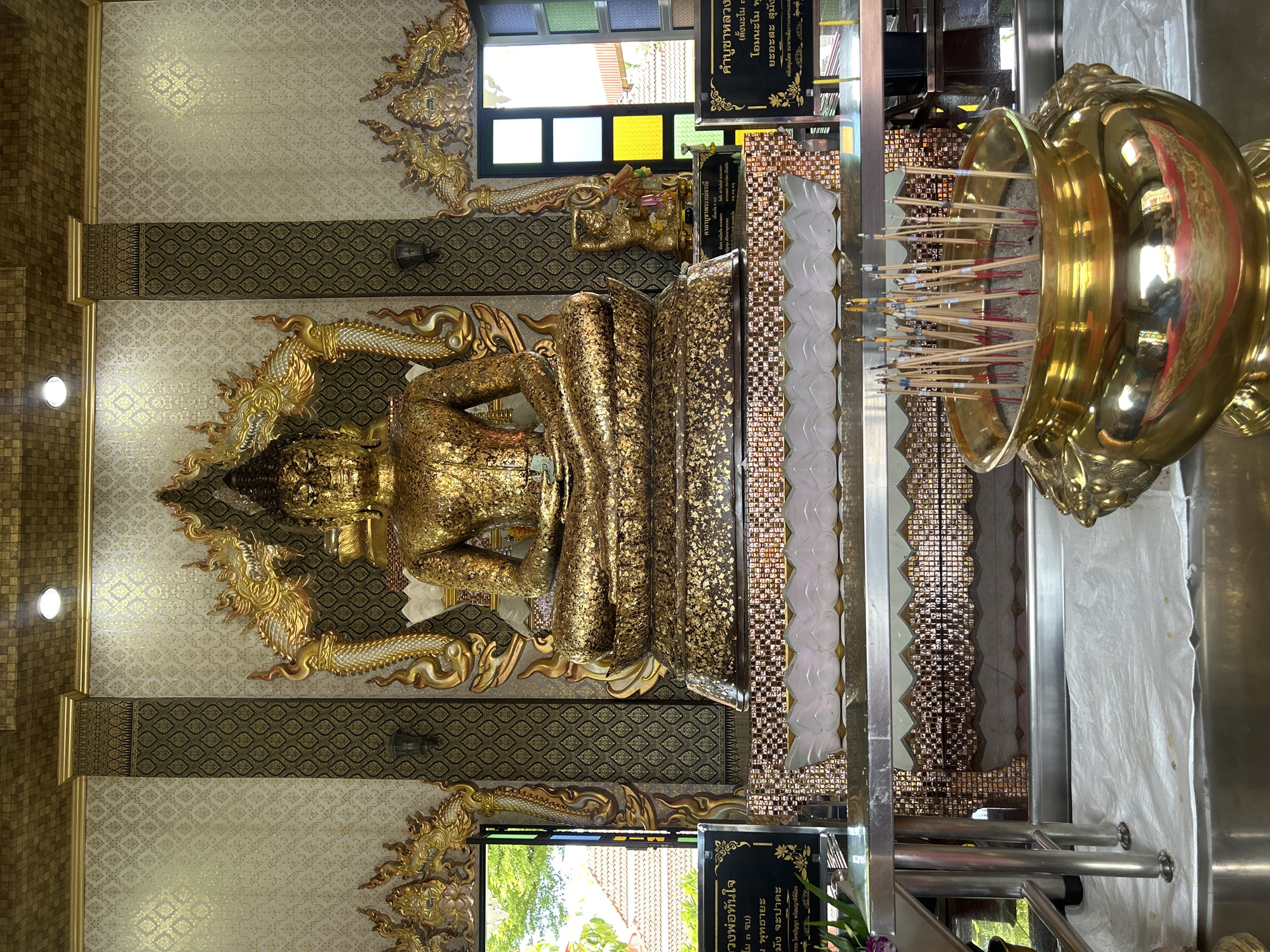 The image shows a large, golden Buddha statue seated in a meditative pose. The statue is adorned with gold leaf. The Buddha is positioned in the center of a room with ornate, gold-colored decorations on the walls and ceiling. There are also several smaller golden statues and decorative elements around the main Buddha. In front of the Buddha, there is a large, golden urn filled with incense sticks. There are also black signs with Thai writing on either side of the urn. The room appears to be a temple or a shrine, likely in Thai Ko Subdistrict, Thailand, given the context. The overall impression is one of reverence and grandeur.