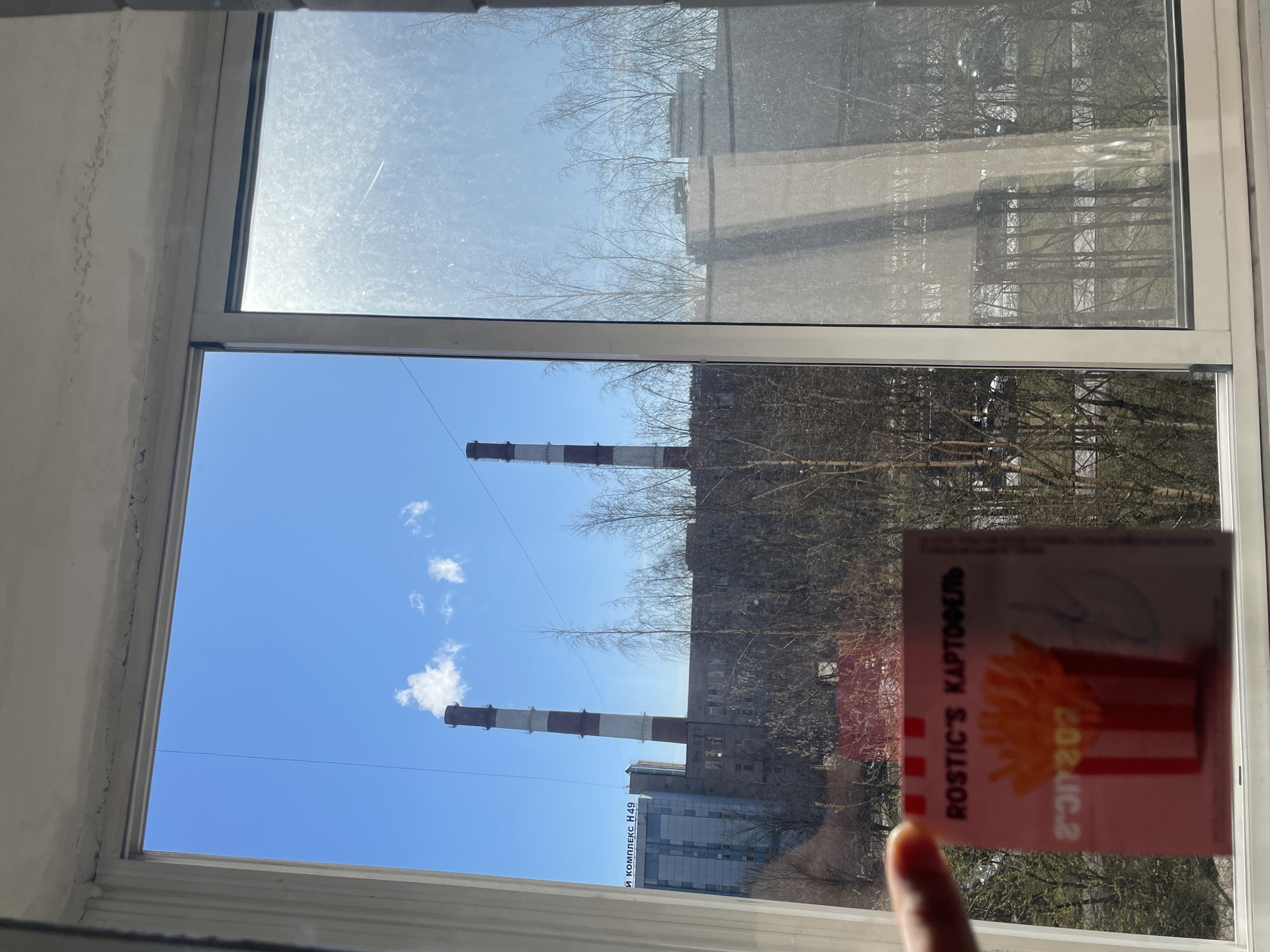 A photograph taken indoors through a window shows a clear blue sky with a few small white clouds, and two smokestacks with alternating red and white bands. A building with the inscription "И КОМПЛЕКС №49" is visible near the base of the smokestacks. A sticker with the inscription "ROSTIC'S КАРТОФЕЛЬ" and an illustration of french fries is partially obstructing the view through the window, with a finger visible at the bottom of the sticker. The location appears to be in the city of Saint Petersburg, Russia, based on the Cyrillic text. The photo is taken through a double-pane window with some streaks or smudges on the glass.