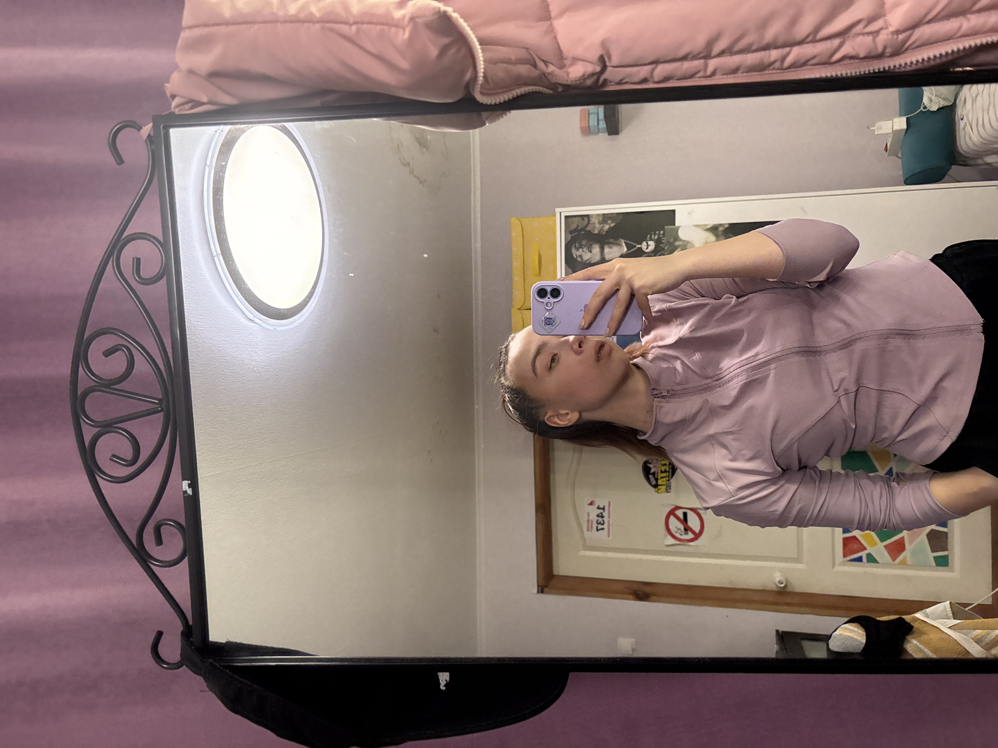 This rotated indoor mirror selfie captures a young woman, likely in her late teens or early twenties, holding a light purple smartphone with a pop socket. She is wearing a lavender long-sleeved athletic jacket and looks directly at her phone with a neutral expression.
The mirror features an ornate black metal frame with scrolled designs on its left side and a small storage area at the bottom. Reflected in the mirror is an interior setting, likely a bedroom. A pink quilted duvet is visible above the mirror, suggesting a bed. To the left of the woman, a circular mirror or light fixture is mounted on the wall.
Behind the woman, a light-colored wooden door with a brown frame is prominent. On the door, there is a framed poster depicting a person with dreadlocks, and a rectangular panel with a colorful geometric pattern, resembling stained glass, on the lower part. Several stickers adorn the door, including a red circular "no smoking" symbol and a white rectangular sticker with the Cyrillic text "ТЕЛ" (TEL) printed vertically. A blue pillow is partially visible at the top right of the reflection, and on the floor, a striped sock and a light-colored shoe are present. The lighting suggests it is daytime indoors. The Cyrillic text "ТЕЛ" serves as a location cue, consistent with Прикамский, Russia.