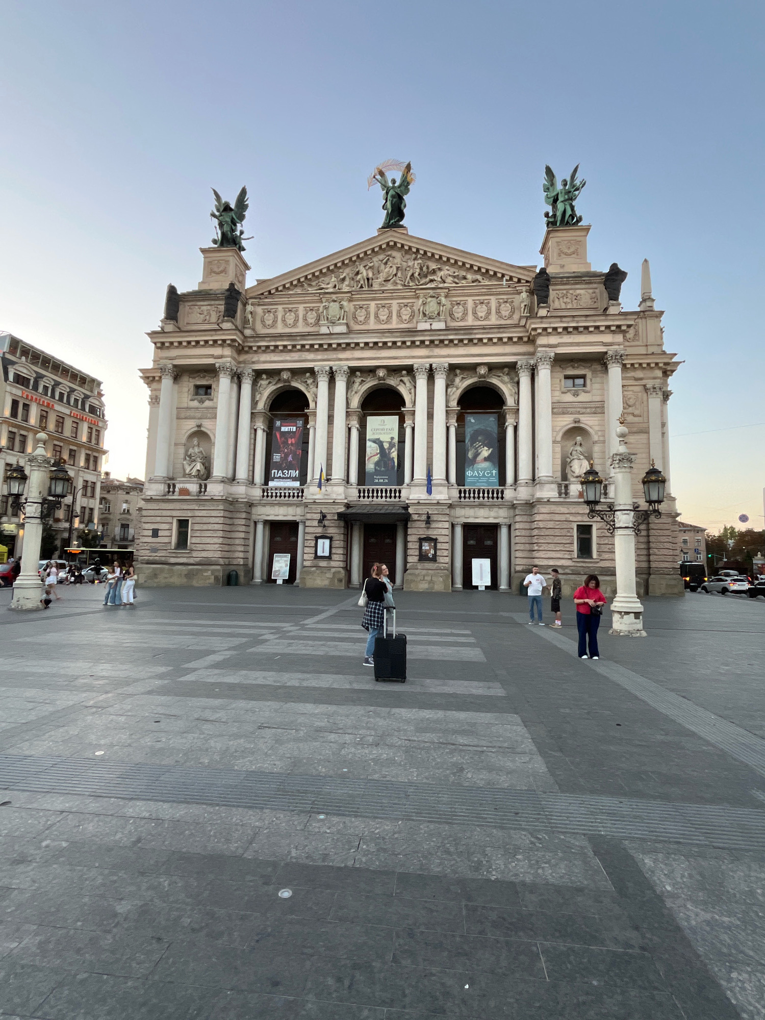 The image depicts the Lviv Opera House, a prominent landmark located in Lviv, Ukraine. The facade of the building is grand, adorned with classical architectural elements, including columns, sculptures of winged figures on the roofline, and intricate carvings. Two large banners are displayed on the front of the building, one depicting "MOTO" and "PAZLI," and another showing the word "FAUST" along with a date.
In the foreground, a paved public square stretches out. Several individuals are present in the scene. A person stands centrally, facing away from the camera, with a black suitcase by their side, appearing to be waiting or preparing to travel. To the right of this person, two individuals are standing, one in a red top and dark trousers, looking down at their phone, and another in a white shirt and dark trousers. Further to the right, a person in a plaid shirt is visible. To the left of the central figure, a group of three individuals are walking together. In the background, a multi-story building with many windows and a distinct architectural style is visible, along with a street with moving vehicles and streetlights. The sky is clear and exhibits a pale blue hue, suggesting either early morning or late afternoon light.