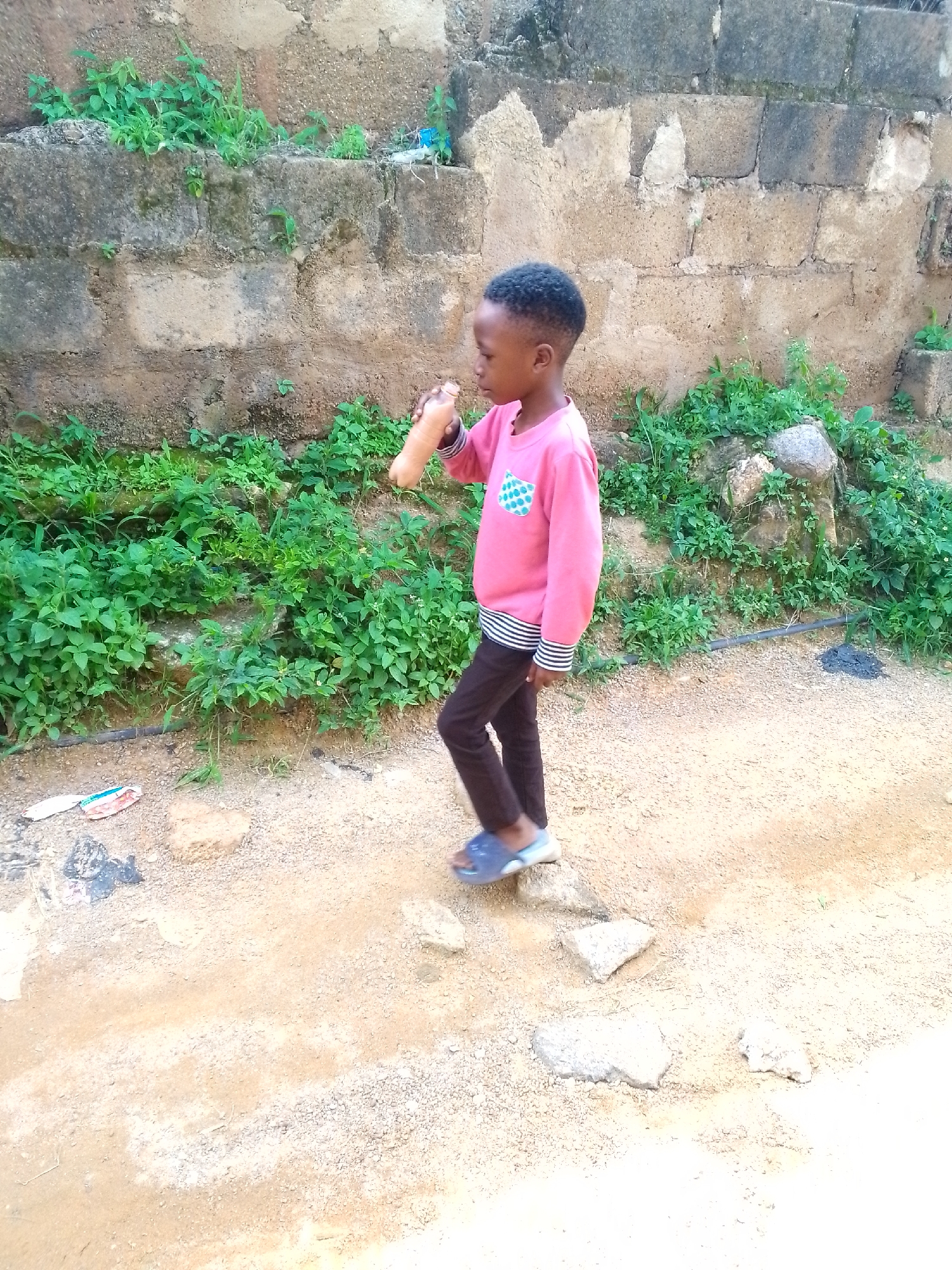 A full-body, side profile shot shows a young Black child, appearing to be a boy, wearing a pink long-sleeved shirt with a green and white patterned pocket on the left side of the chest and black pants with a striped band at the wrists and ankles, walking on a dirt path. The child is holding a light brown object, possibly a bottle, up to their mouth with their right hand. The child is wearing blue sandals. The child is walking in front of a rough, tan-colored brick wall covered in moss and overgrowth. To the left of the child, a patch of green vegetation is visible, and to the right, more dense green plants and some rocks are present. The ground is composed of dirt and gravel with a few scattered stones and some debris, including what appears to be trash. The lighting suggests it is daytime.