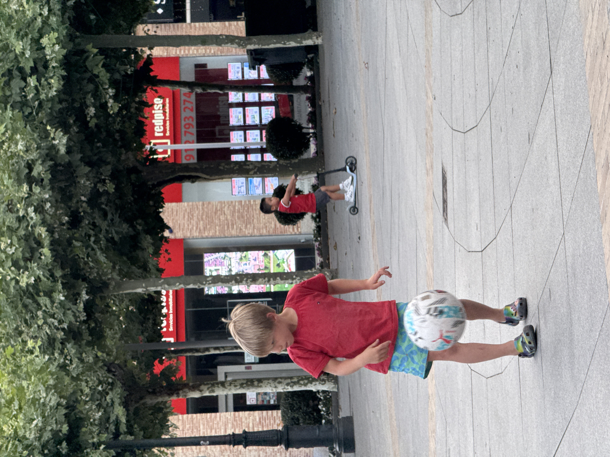 The image depicts a boy in a red t-shirt and patterned shorts performing a soccer trick outdoors on a paved walkway. He is in the process of kicking a soccer ball, which is blurred due to motion. His left foot is raised and positioned behind the ball, while his right foot is on the ground, supporting his balance. His expression is focused.
In the background, another child is visible riding a scooter. Further back, buildings with large glass windows display what appear to be advertisements or listings, with the text "redpise Servicios Inmobiliarios" and a phone number "912 793 274" clearly visible on a red sign. Trees with green foliage line the walkway, suggesting a pleasant outdoor environment, possibly during daylight hours. The pavement is made of large, light-colored tiles. The overall scene suggests a casual recreational activity in an urban or commercial setting.