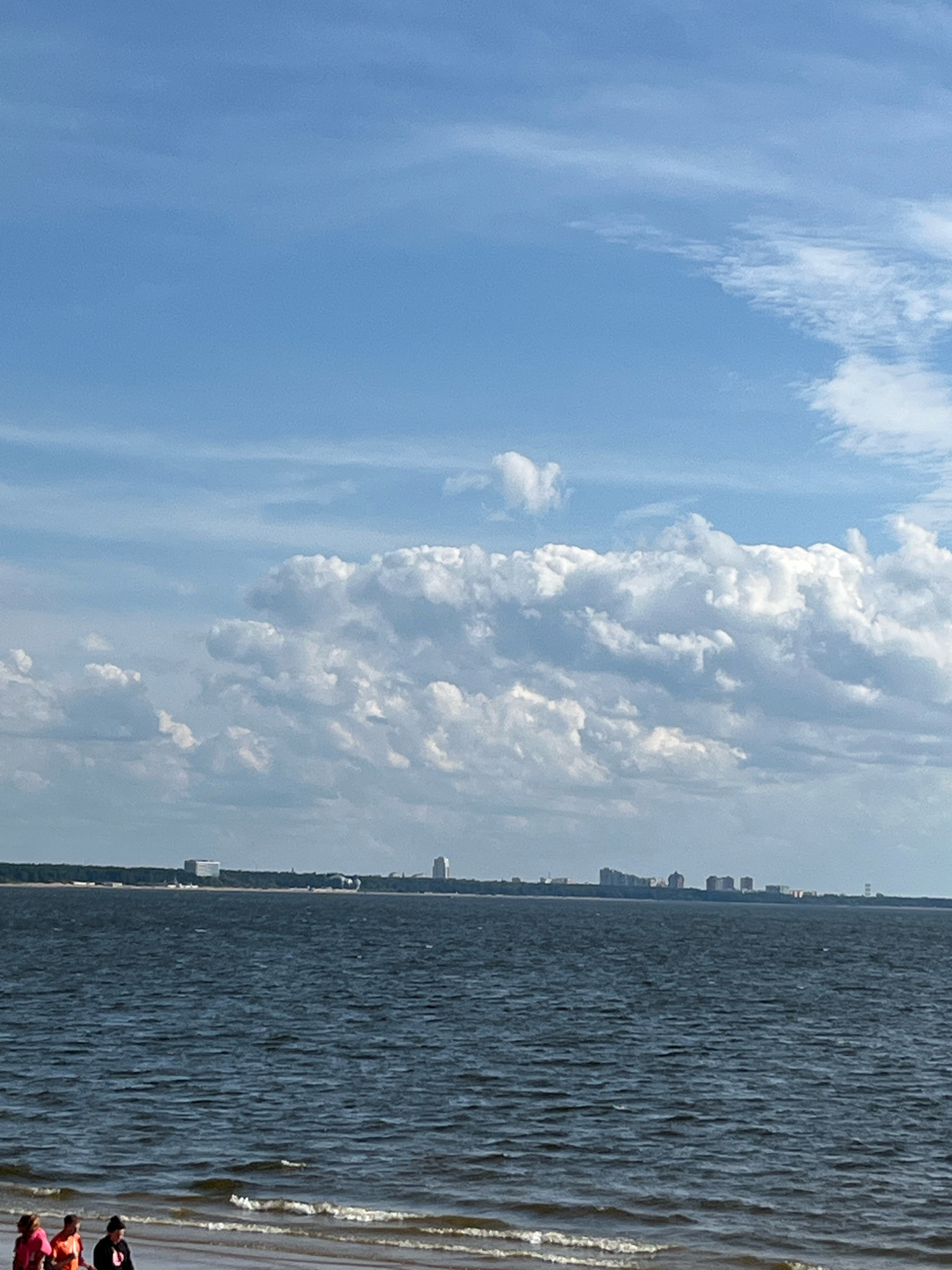 The image captures a wide-angle view of a body of water, likely a gulf or sea, with a city skyline in the distance. The foreground features a sandy beach with a few people walking along the water's edge. The water is dark and rippled, suggesting a breezy day. The sky above is a bright blue, dotted with large, fluffy white clouds. The city skyline, visible across the water, consists of several buildings of varying heights, suggesting an urban landscape. The overall impression is one of a serene, possibly coastal, environment. Given the prompt's reference to Solnechnoye, Russia, it's plausible this scene depicts a location along the Gulf of Finland near that area.