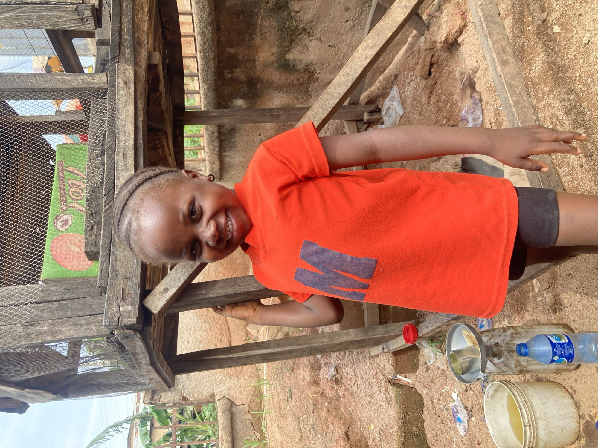A young Black girl with braided hair and a dark earring stands outdoors, smiling at the camera. She is wearing a bright orange t-shirt with a dark "M" on the front, and black shorts. She is standing in front of a rustic, multi-tiered wooden structure. The ground is covered in reddish-brown dirt and small pebbles, with some scattered plastic debris. To the girl's left, on the ground, are a white bucket containing a yellowish liquid, a blue water bottle, and a metal funnel with a can inside, resting on top of a plastic container. The background shows a rough concrete wall and some greenery. The overall setting appears to be a casual, possibly residential or commercial outdoor area in Gwong, Nigeria.