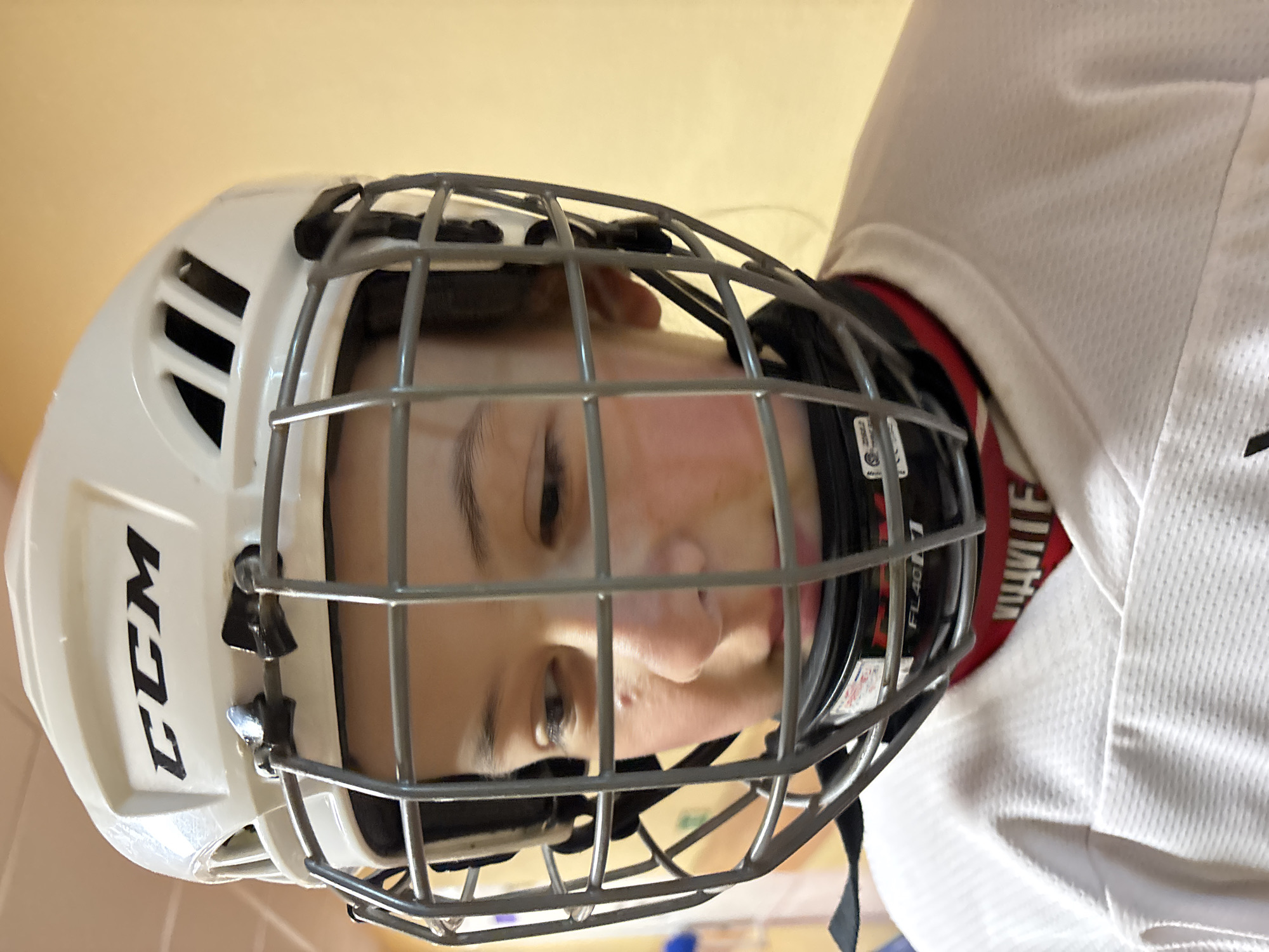 A person wearing a white hockey helmet with a gray metal face mask and a white jersey is positioned indoors. The helmet has "CCM" printed on the side. A red collar is visible beneath the jersey. The location is specified as the city of Chaikovsky, Russia, although this detail is not discernible from the visual information in the image.