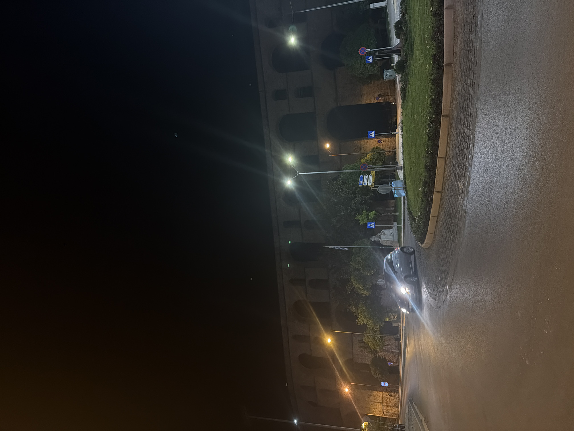 Night scene in Kavala, Greece. The historic Kamares aqueduct is visible, illuminated by streetlights. A car is driving on the wet road alongside a small green area with signage. The sky is dark.