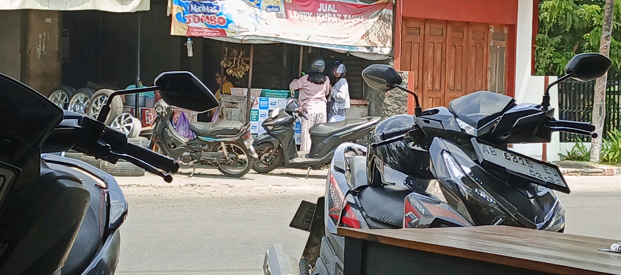 The image shows a street scene in Godean, Indonesia, with several motorcycles parked in front of a shop. The shop has a banner advertising "Mari Mas Jumbo" and "JUALOTEK KUPAT TAHU". There are three people visible, two women wearing headscarves and one person in the background. One woman is standing next to a scooter, wearing a pink top and a long skirt. The other woman is wearing a white shirt and blue pants. In the foreground, there are two motorcycles, one black and one silver, with parts and accessories scattered around. The environment appears to be a typical Indonesian street scene, with a building in the background and lush green foliage. The overall impression is one of a bustling local market or commercial area.