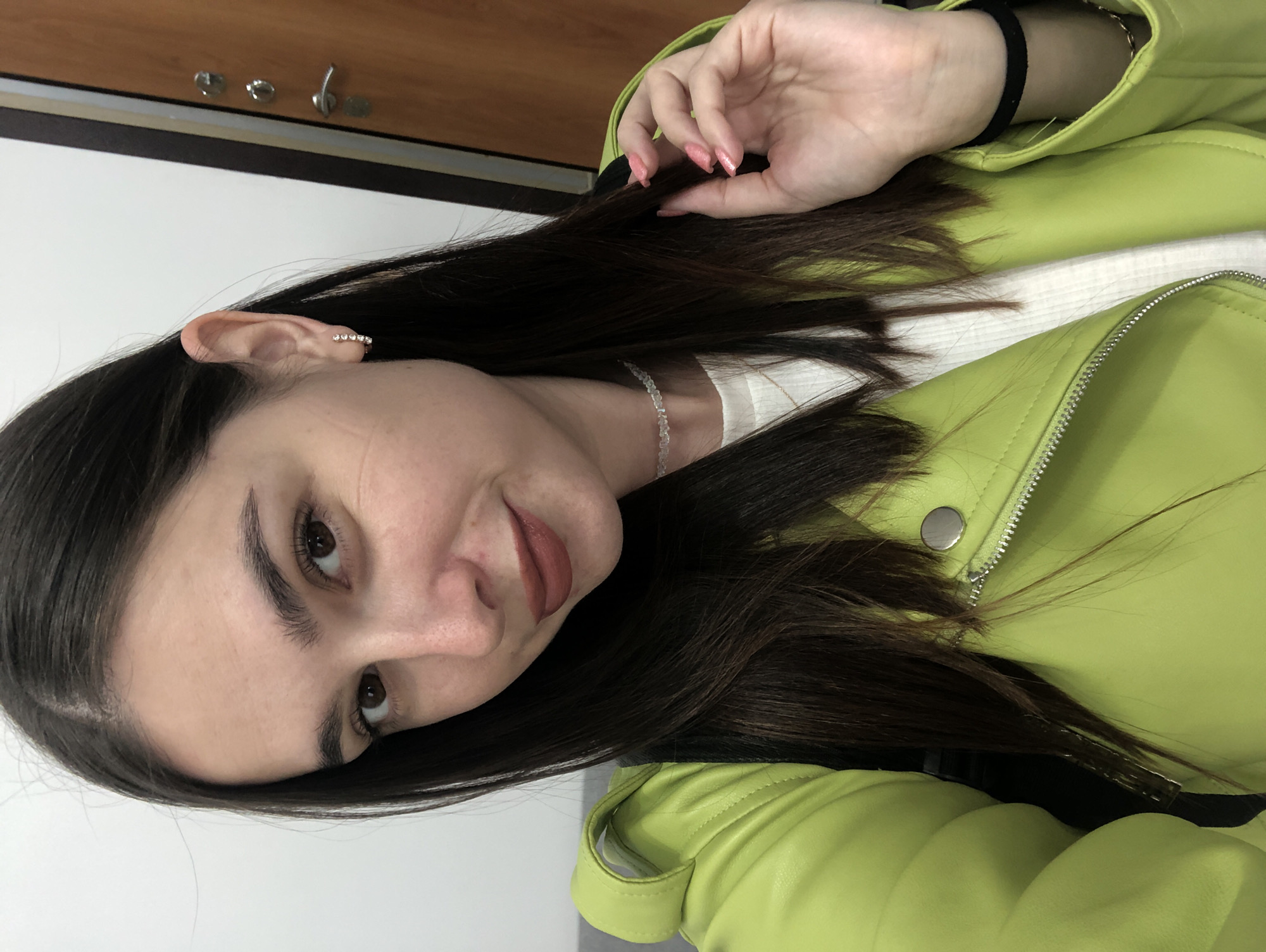 This is a portrait of a young woman, likely taken in Chaikovsky, Russia, given the provided location context. The woman has long, dark hair, which is partially obscuring her face and is styled over her shoulder. She is wearing a bright, lime green jacket with a zipper and button details, over a white top. A thin, beaded necklace is visible around her neck. She is also wearing a stud earring in her visible ear and a black band on her wrist. Her right hand is resting on her hair, near her head, and her fingernails are painted a light pink. The background appears to be indoors, with a light-colored wall and what looks like a wooden surface with some hardware at the top. The lighting suggests it is daytime, and there are no obvious indicators of weather. The woman's expression is neutral, and she appears to be posing for the camera. The image is taken from a close-up perspective, focusing on the woman's face, upper body, and accessories. There is no visible text in the image.