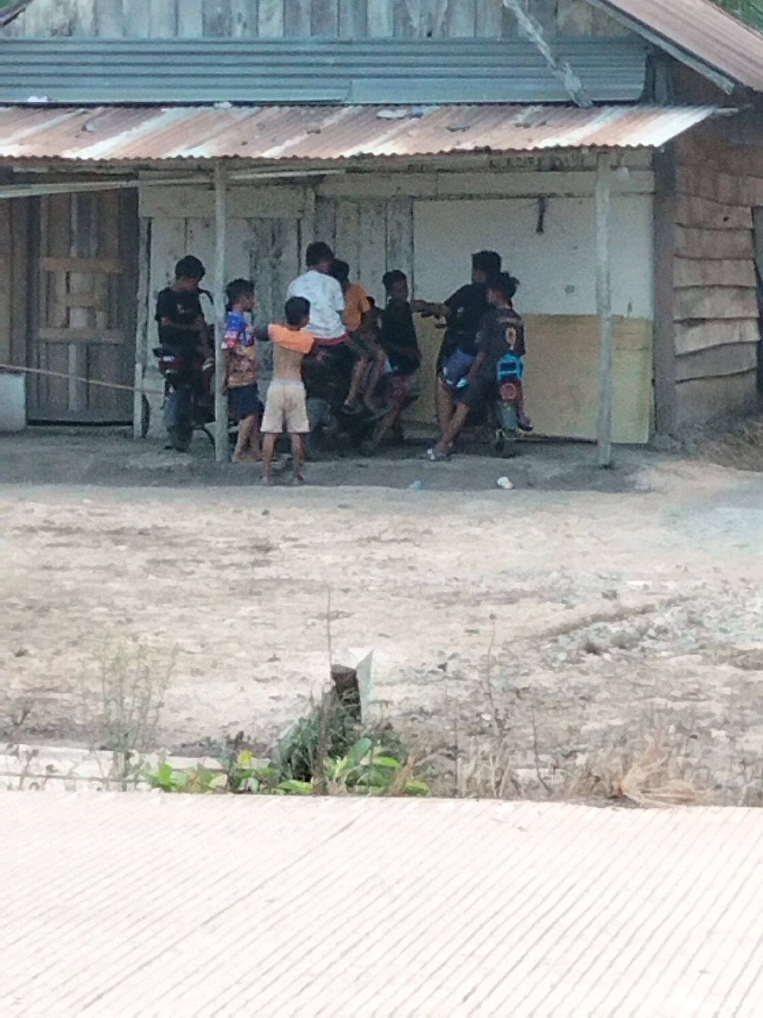 In Perambahan, Indonesia, a group of young people are gathered on and around two motorcycles in front of a weathered, open-fronted structure with a corrugated metal roof. The structure appears to be a makeshift shelter or shop. The ground is dirt and gravel, with sparse vegetation. The individuals are dressed casually, and some are sitting on the motorcycles while others stand. One person in the foreground is wearing an orange t-shirt and light-colored shorts, facing away from the camera. The scene suggests a casual, everyday moment in a rural or semi-rural setting.