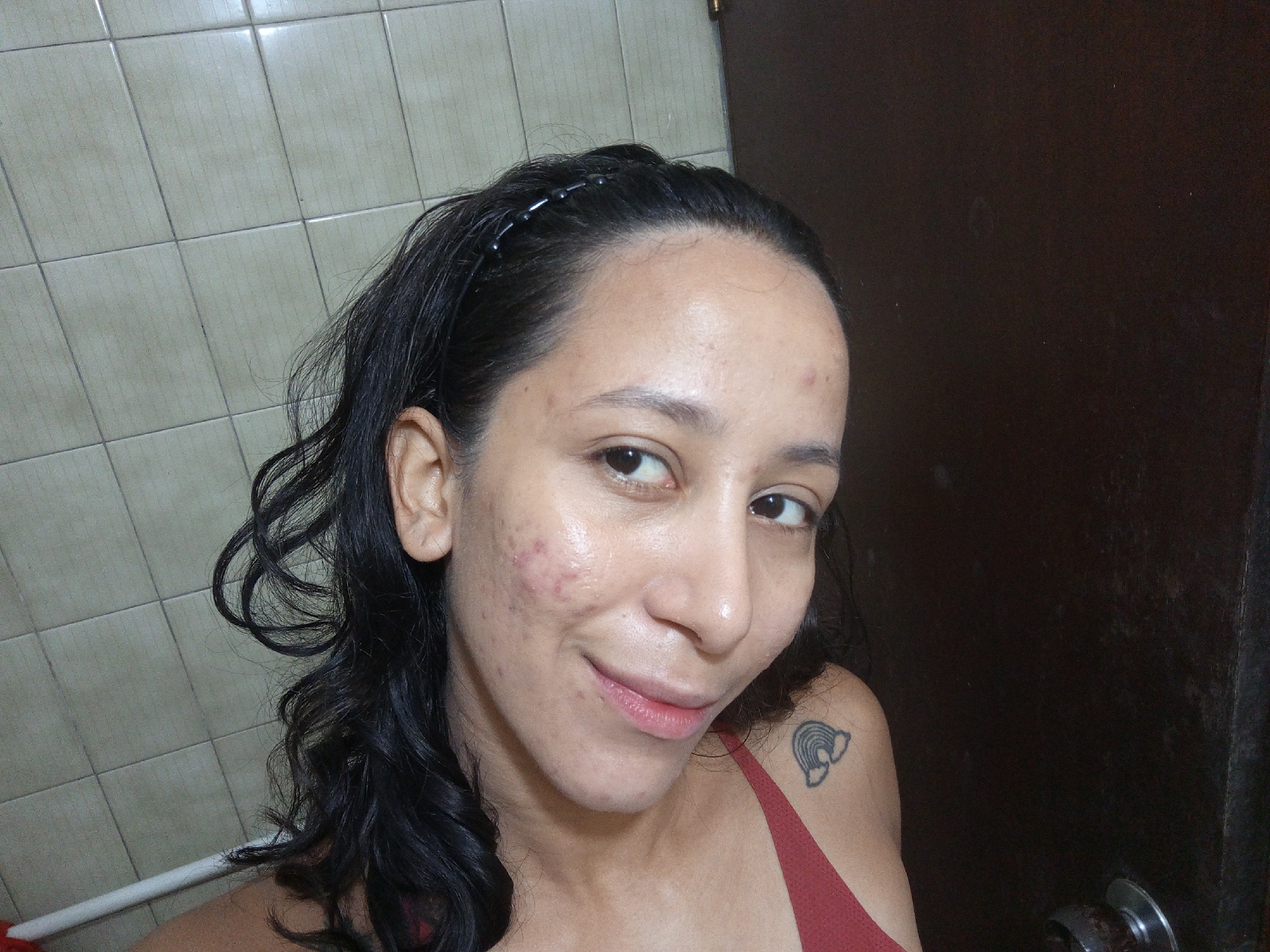 A woman with dark hair pulled back with a beaded headband smiles at the camera. She is wearing a red tank top and has a tattoo of a rainbow with clouds on her left shoulder. Her skin has some acne breakouts. She is standing in a bathroom with beige tile walls and a dark brown door. The location is likely Porlamar, Venezuela.